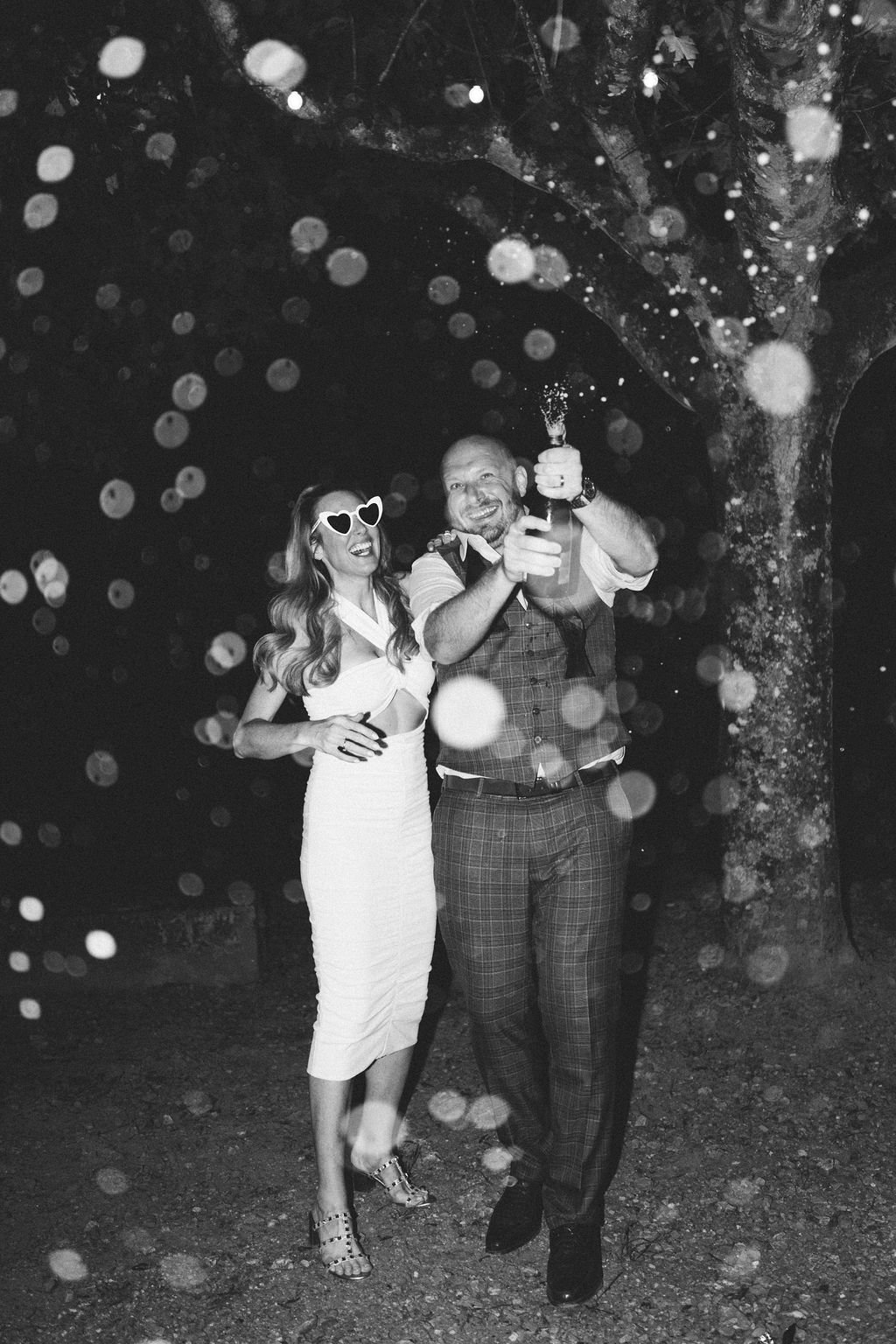  A black &amp; white image of the groom popping a bottle of champagne outside, as the bride is stood next to him wearing heart shaped sunglasses. 