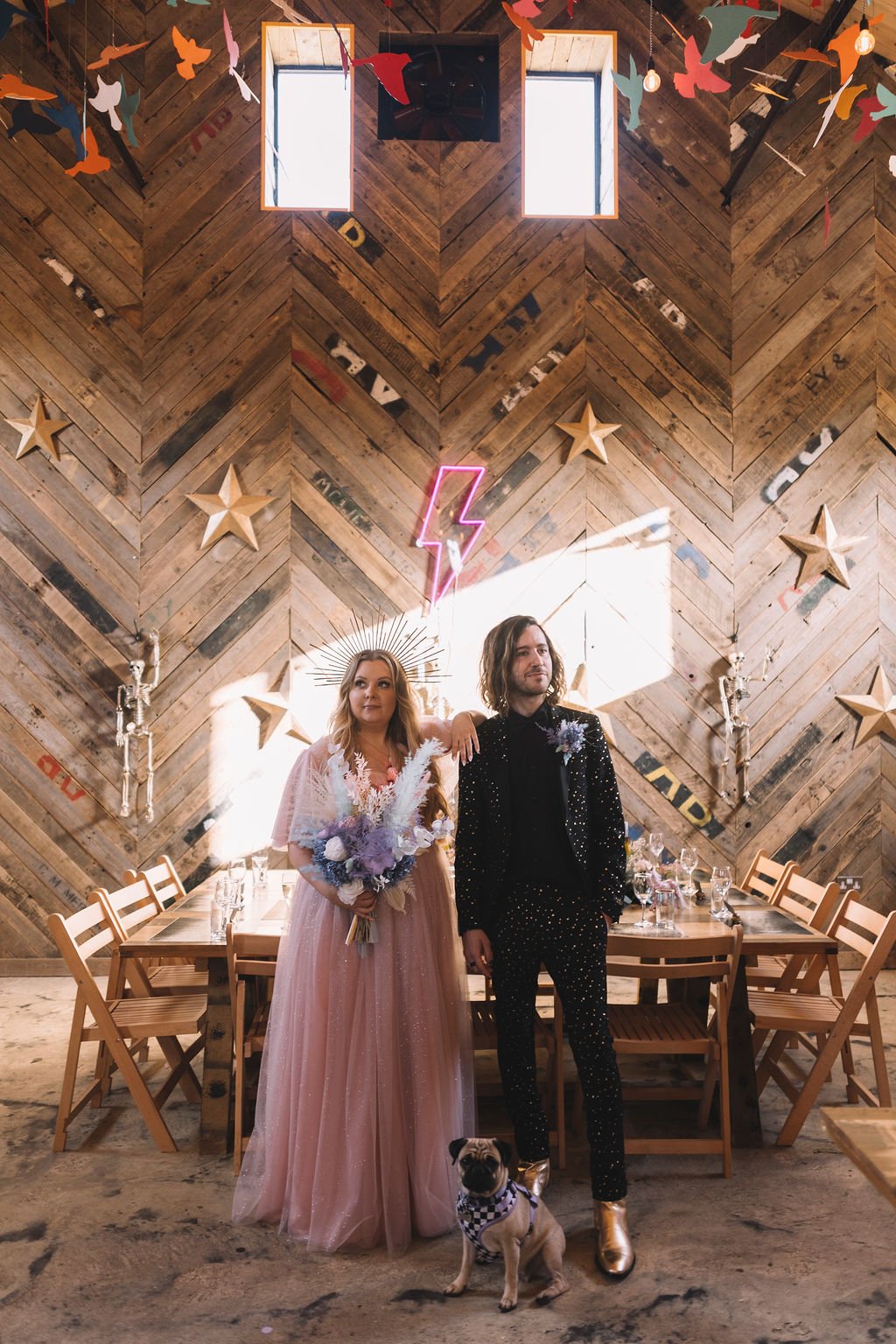  Wedding photographer has taken this image of the bride and groom inside at their wedding venue, The Canary Shed in Essex. the bride is wearing a pink wedding dress and the groom is wearing a black suit covered with stone embellishments.   Behind the