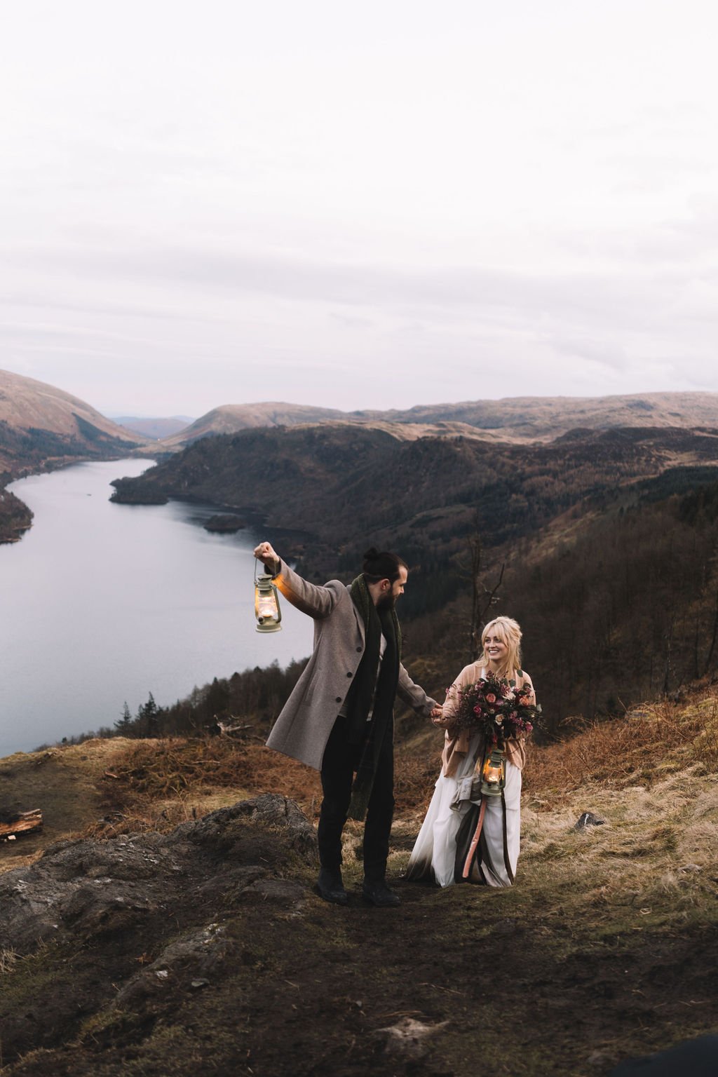  A natural image of a bride and groom as the walk up a hillside wearing their wedding outfits and carrying lit oil lamps. 