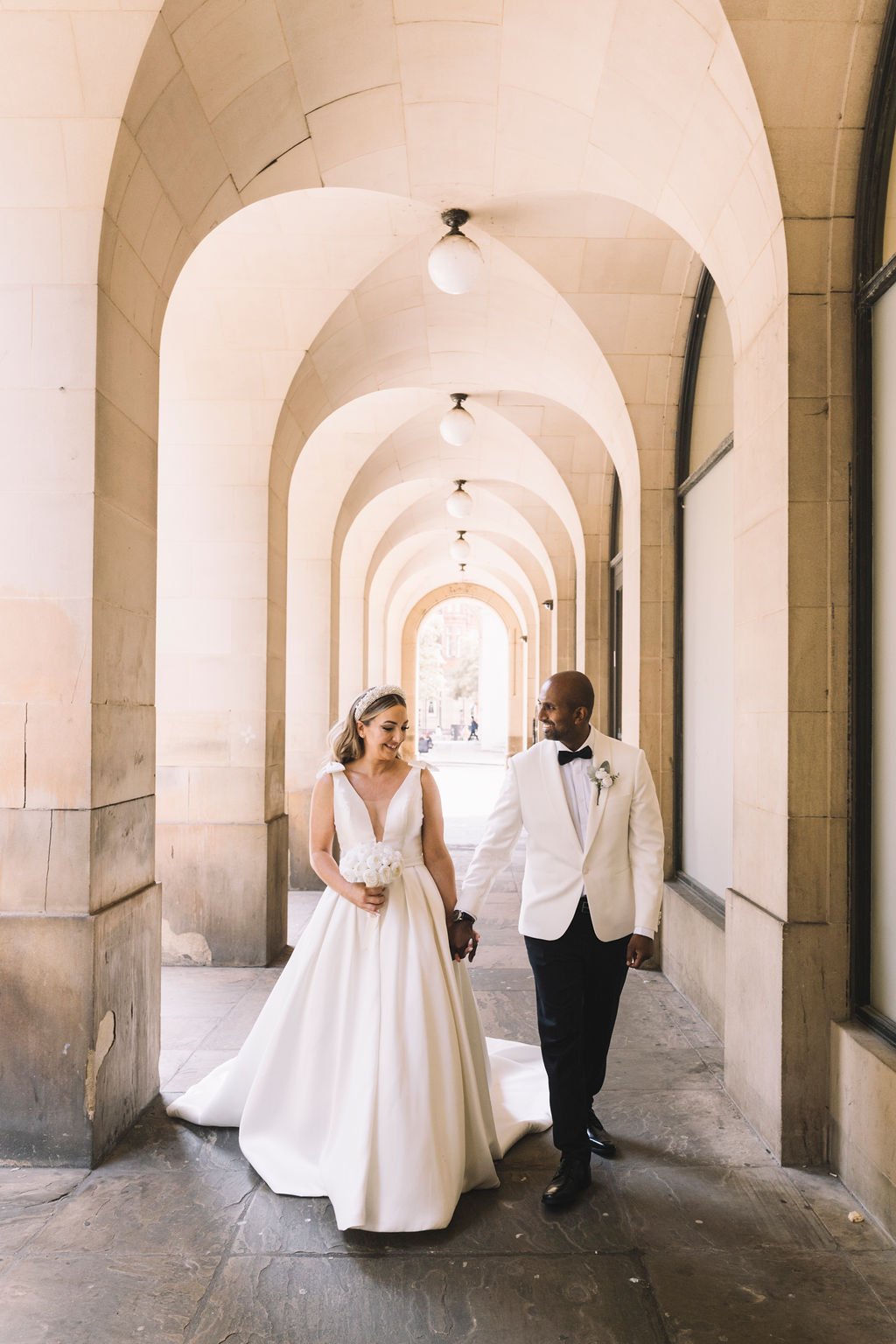  Using the light perfectly and to their advantage, the wedding photographer has captured the bright arches the bride and groom are walking under outside. The newlywed couple are sharing an intimate moment between them as they walk together holding ha