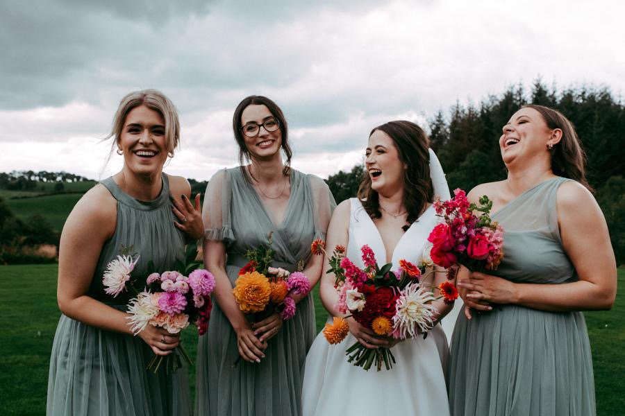  Stunning, modern &amp; bright hand-tied bouquets held by bride and bridesmaids. The wedding party florals bright oranges and pinks, pop against the sage green bridesmaid dresses and the bride’s white dress.  Photo by Pocket Square Photography 