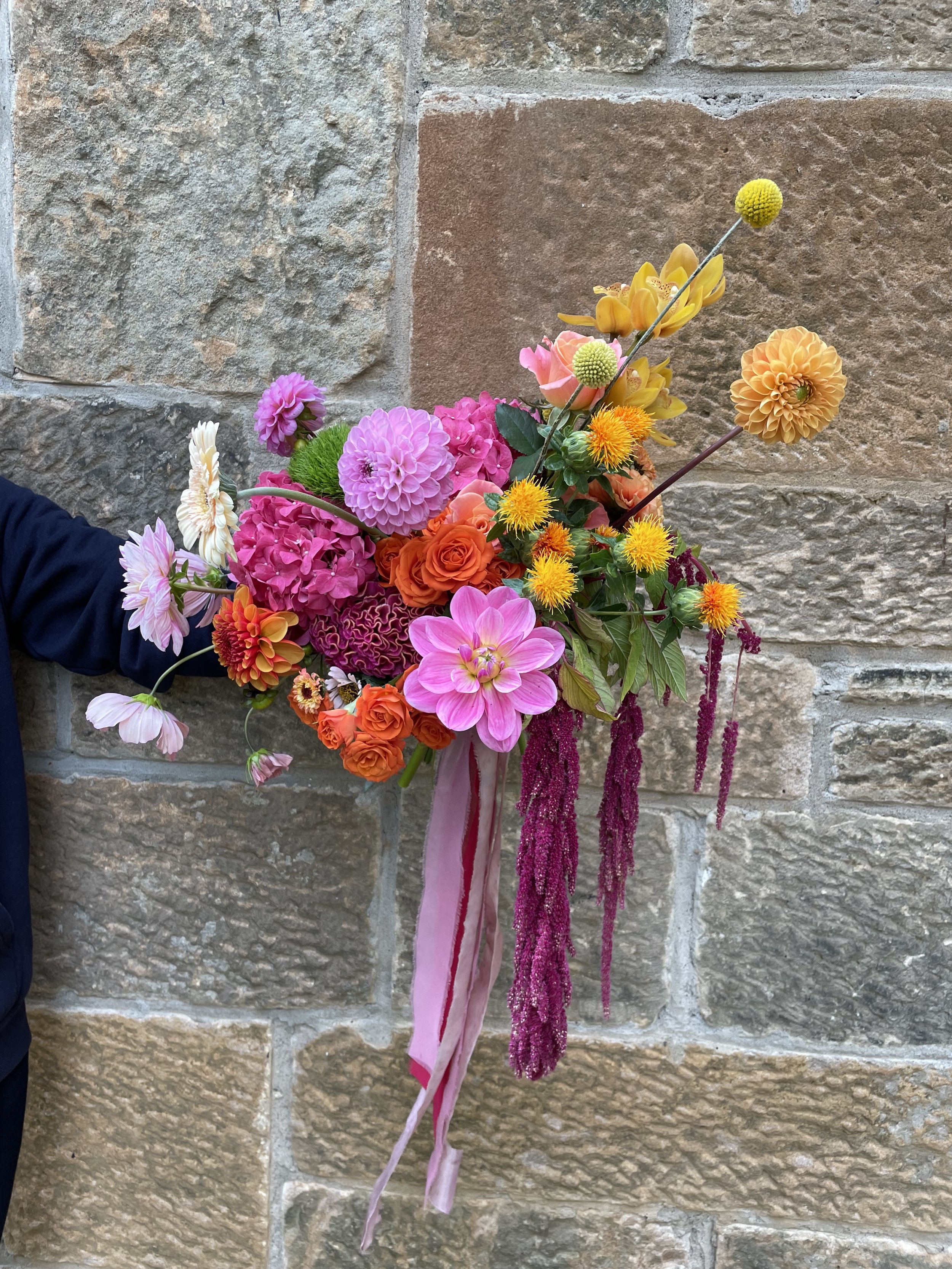  A bright modern wedding bouquet is being held in front of a stone wall. The different styles and textures of the blooms adds lots of fun and interest, as well as the ribbon it has been tied with along with the hanging florals. 
