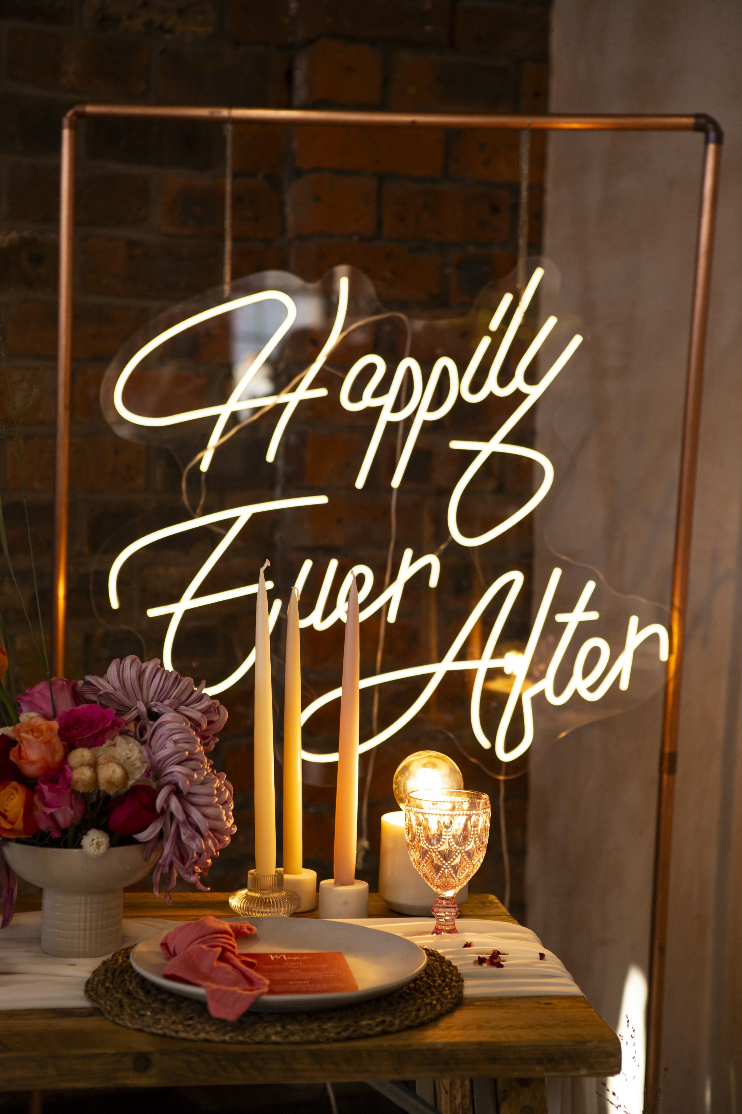  A white neon HAPPILY EVER AFTER sign is displayed inside a copper pipe frame with a gorgeous bowl of modern florals, a lightbulb lamp, 3 white taper candles and a retro wine glass. 