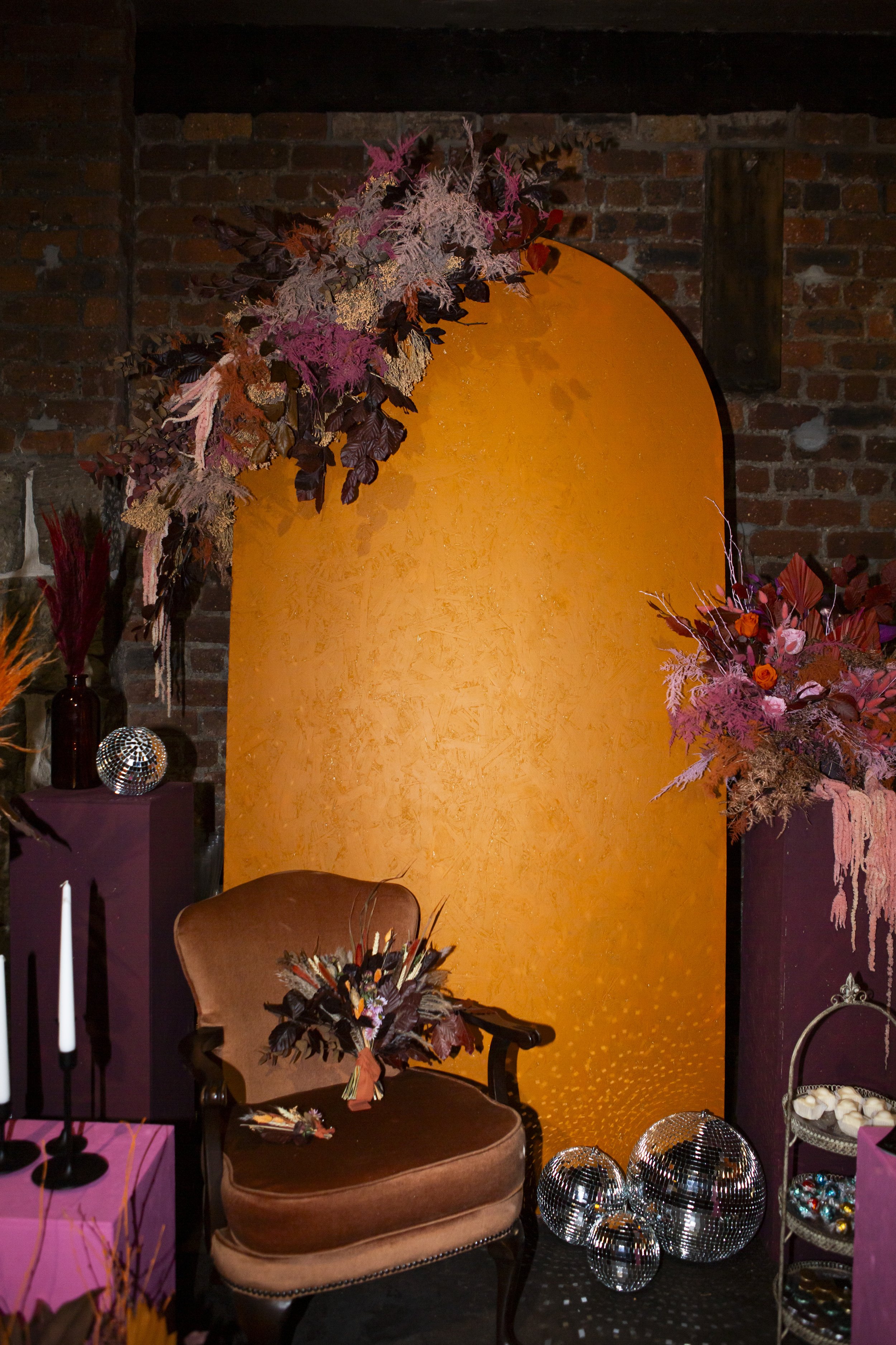  Retro feel styled space. Brown suede armchair, taper candles on pink tablecloth, glass bowls on the floor and various dried floral arrangements. 