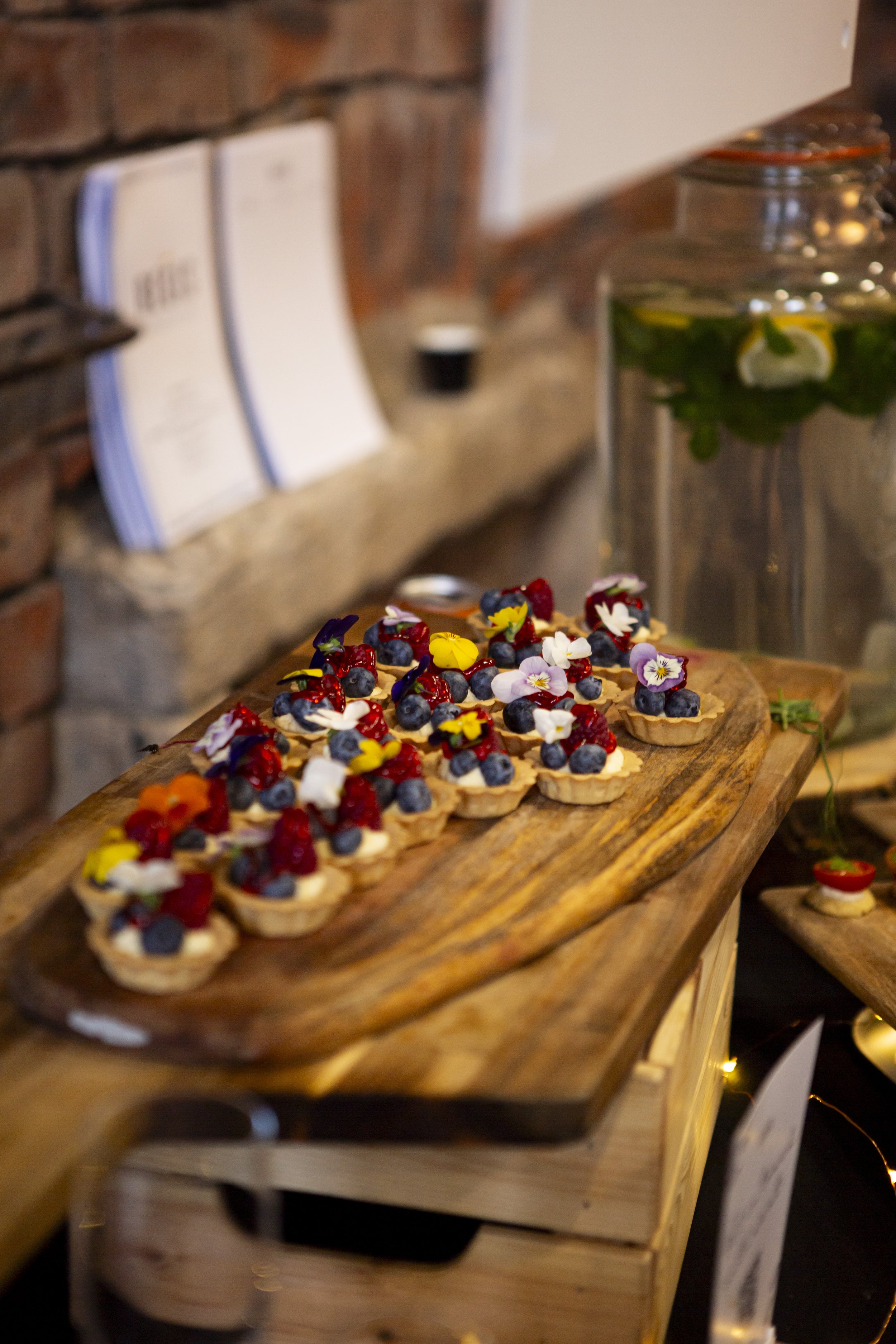  Bite sized pastry cases filled with cream and berries, displayed on a wooden board which has been placed on a wooden unit. 