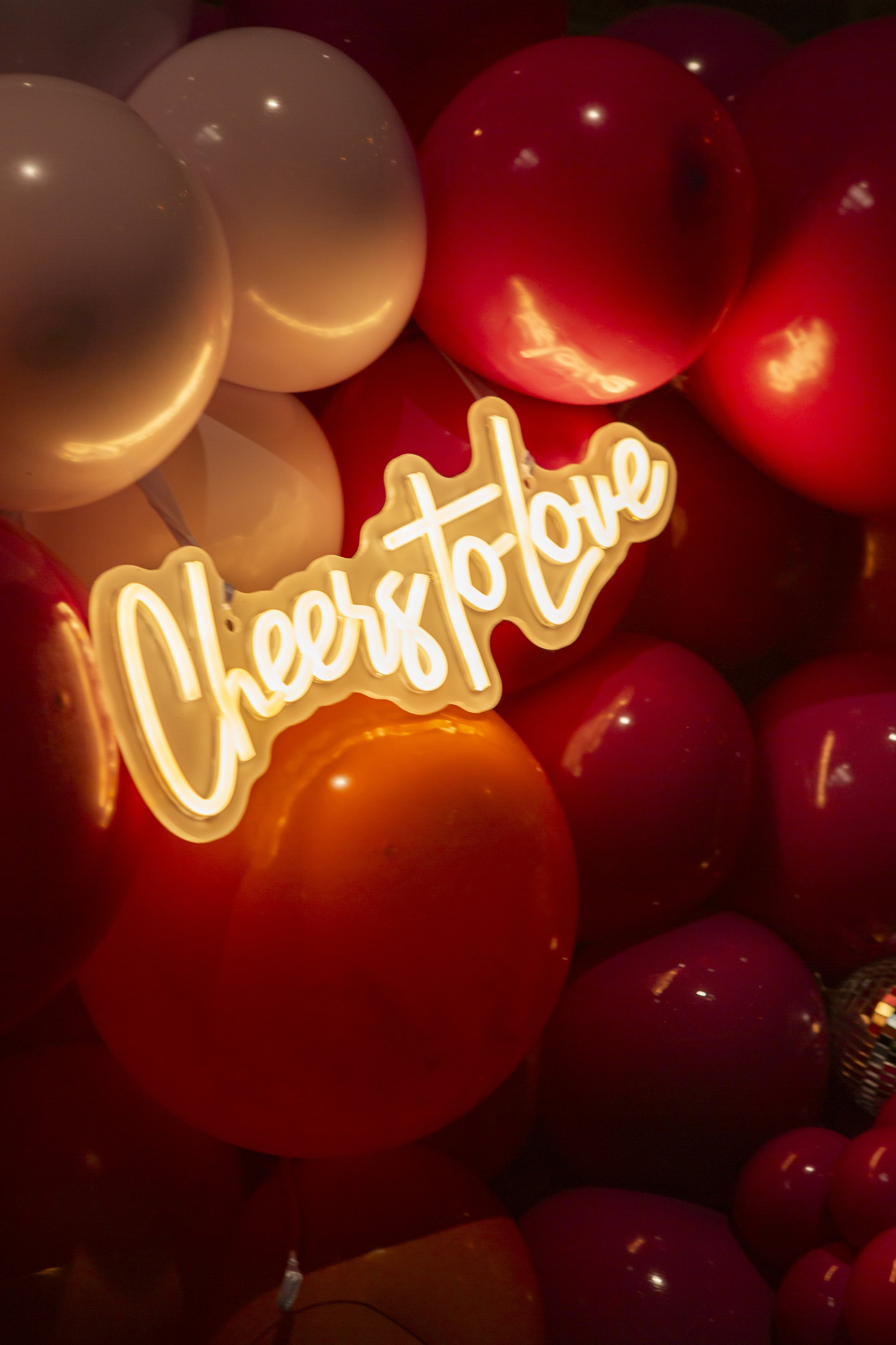  Close up image of white neon CHEERS TO LOVE sign nestled amongst bright orange, red and white balls. 