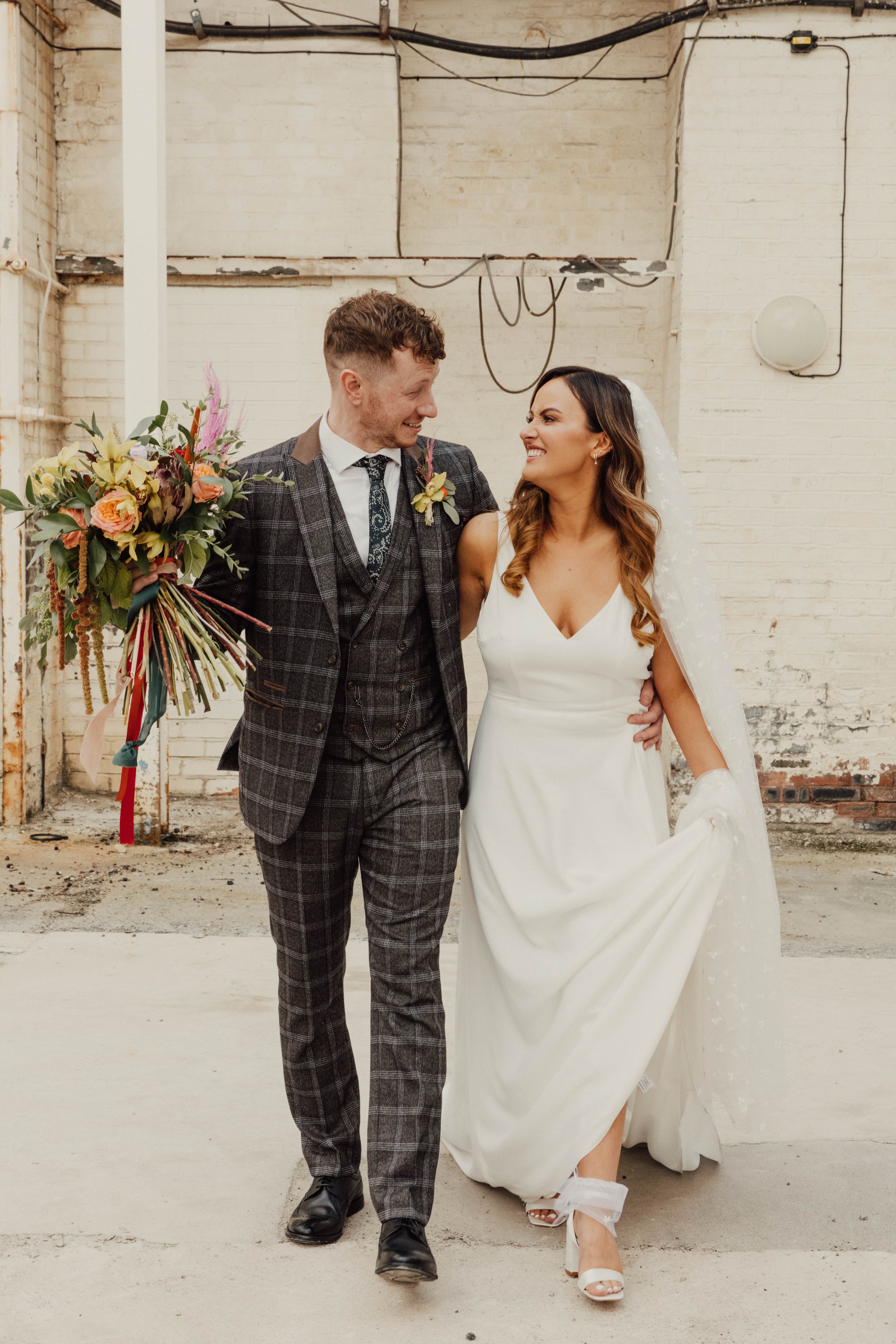  Within a white industrial style building the bride and groom are stood looking at each other with their arms around each other. The groom is wearing a tartan 3 piece suit and is holding the brides bouquet which has been created with bright real, dye