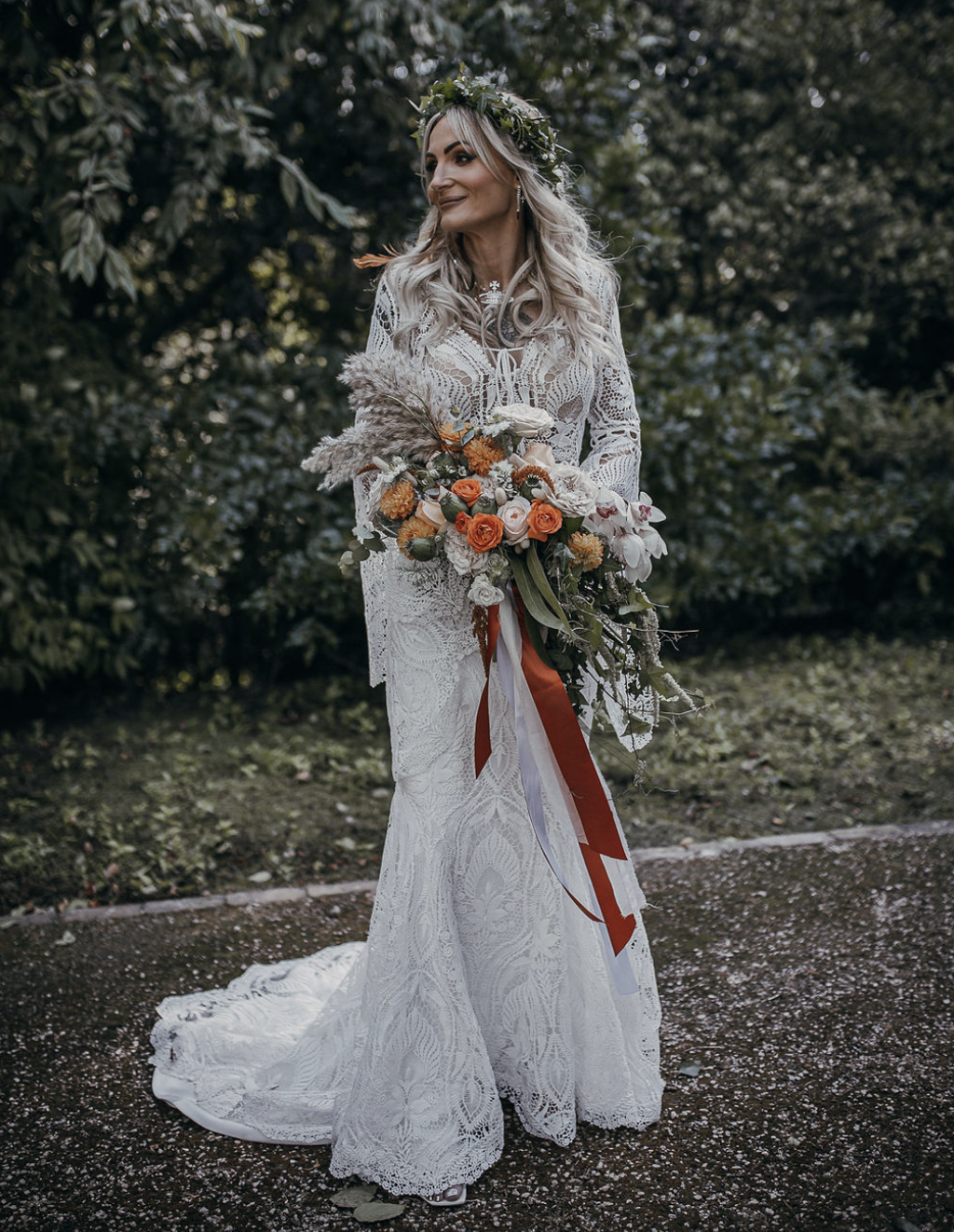  A bride wearing a long sleeved cream wedding dress is stood outside holding an original floral creation, filled with dried and natural flowers. The bridal bouquet is tied with orange and white ribbons to match the flowers. The bride is wearing a flo