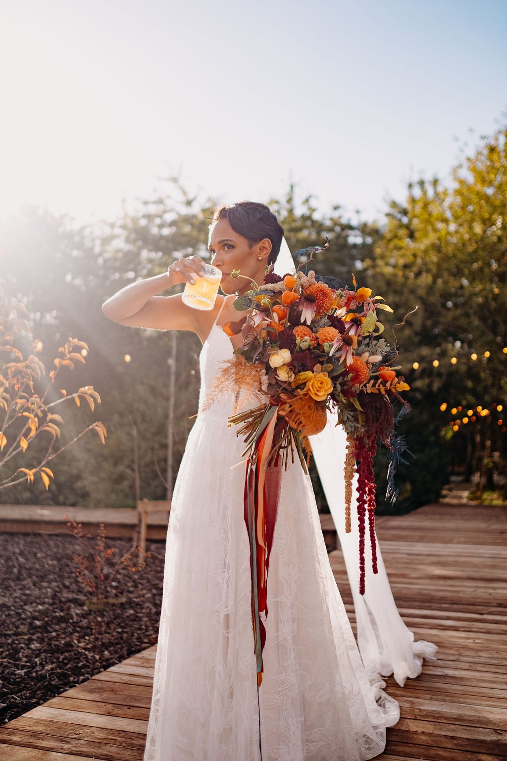  A bride stood outside in the sunshine having a drink whilst holding her epic floral creation. The fun and bright hand-tied bridal bouquet is filled with natural, dyed and dried florals and foliage and the creative design is completed with the quirky