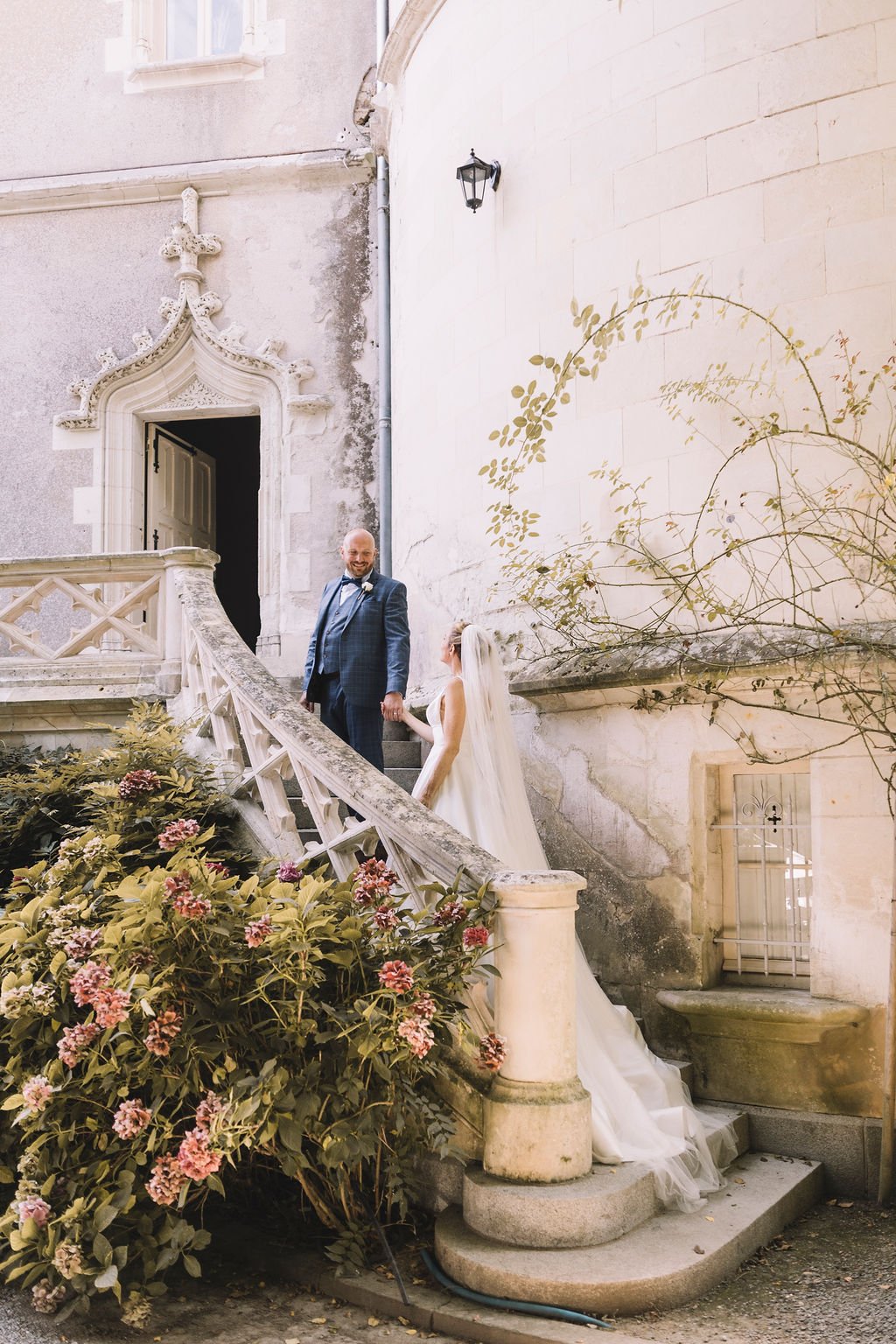 The wedding photographer has captured the moment the bride and groom walk up a beautiful stone staircase outside. The groom is ahead and is looking back towards the bride as they hold hands.  