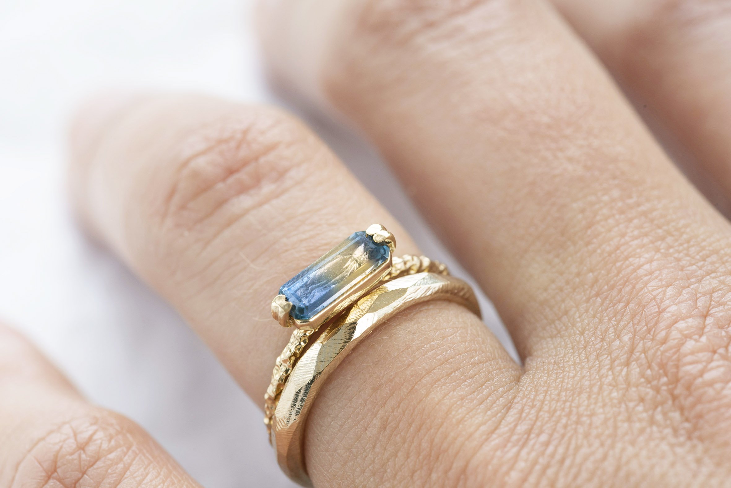  Two wedding rings. A thin gold textured band with modern playful shaped blue stone set in on top and below there is a slim, plain gold band with subtle graphic pattern,  