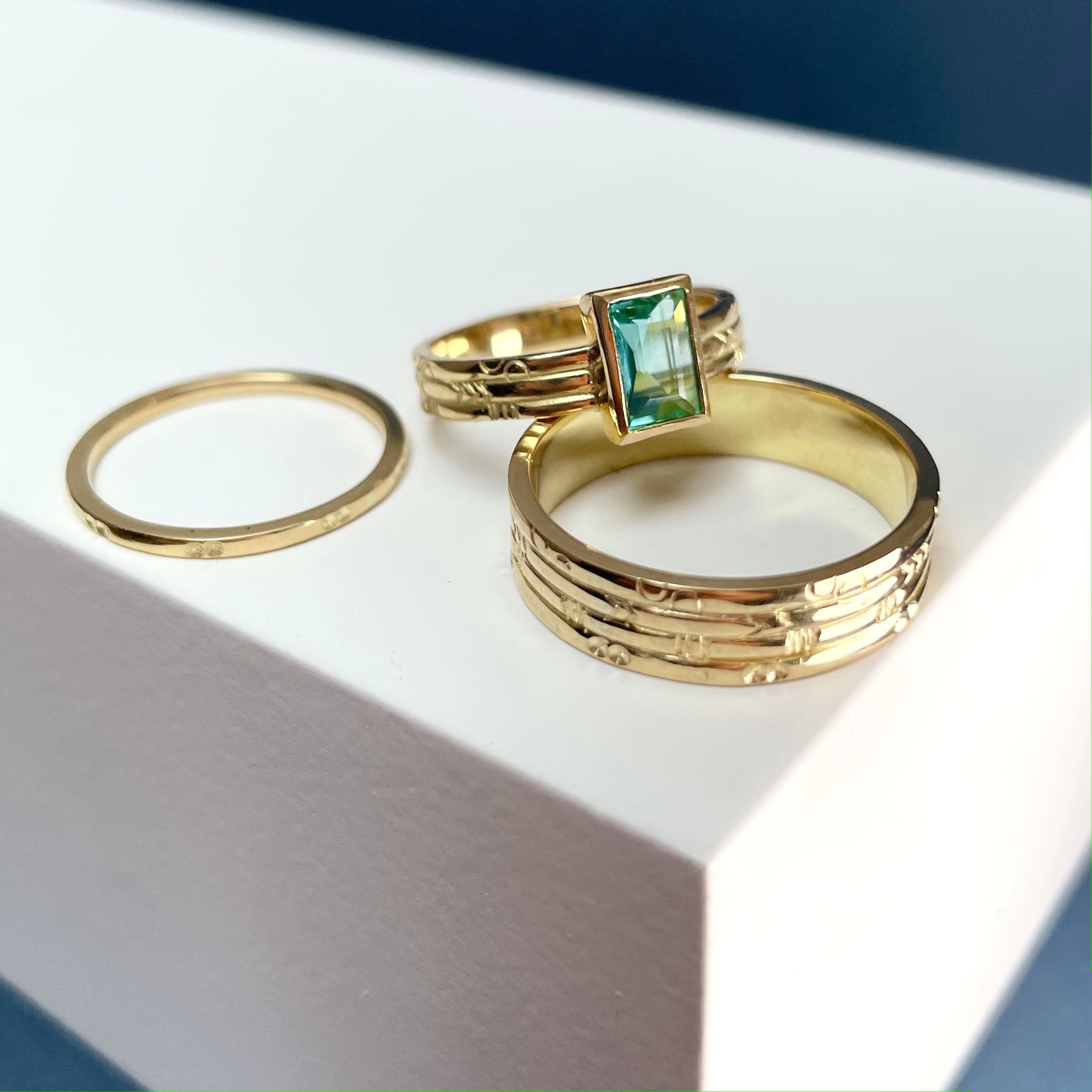  Three rings, two gold wedding bands with subtle graphic detailing in different widths and a gold engagement ring which compliments the wedding bands and in addition has a rectangular green stone set on top. 