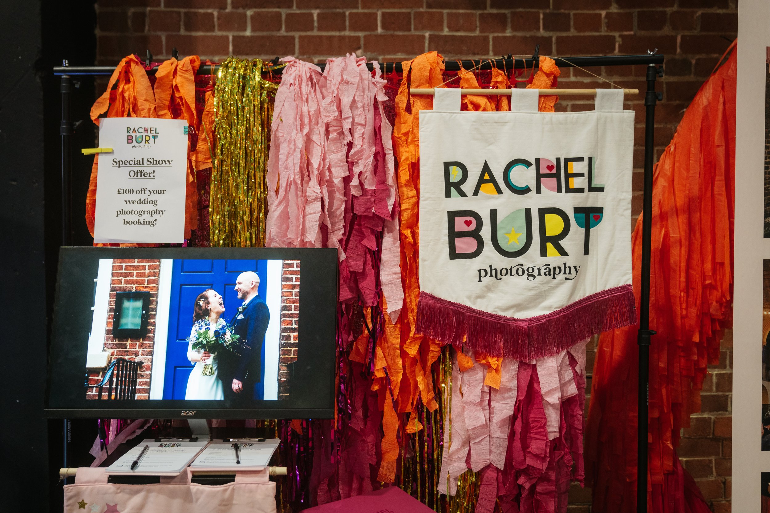  RACHEL BURT PHOTOGRAPHY banner is hung against a backdrop of multicoloured streamers with a monitor in front showcasing some of the incredible wedding photographers work. 