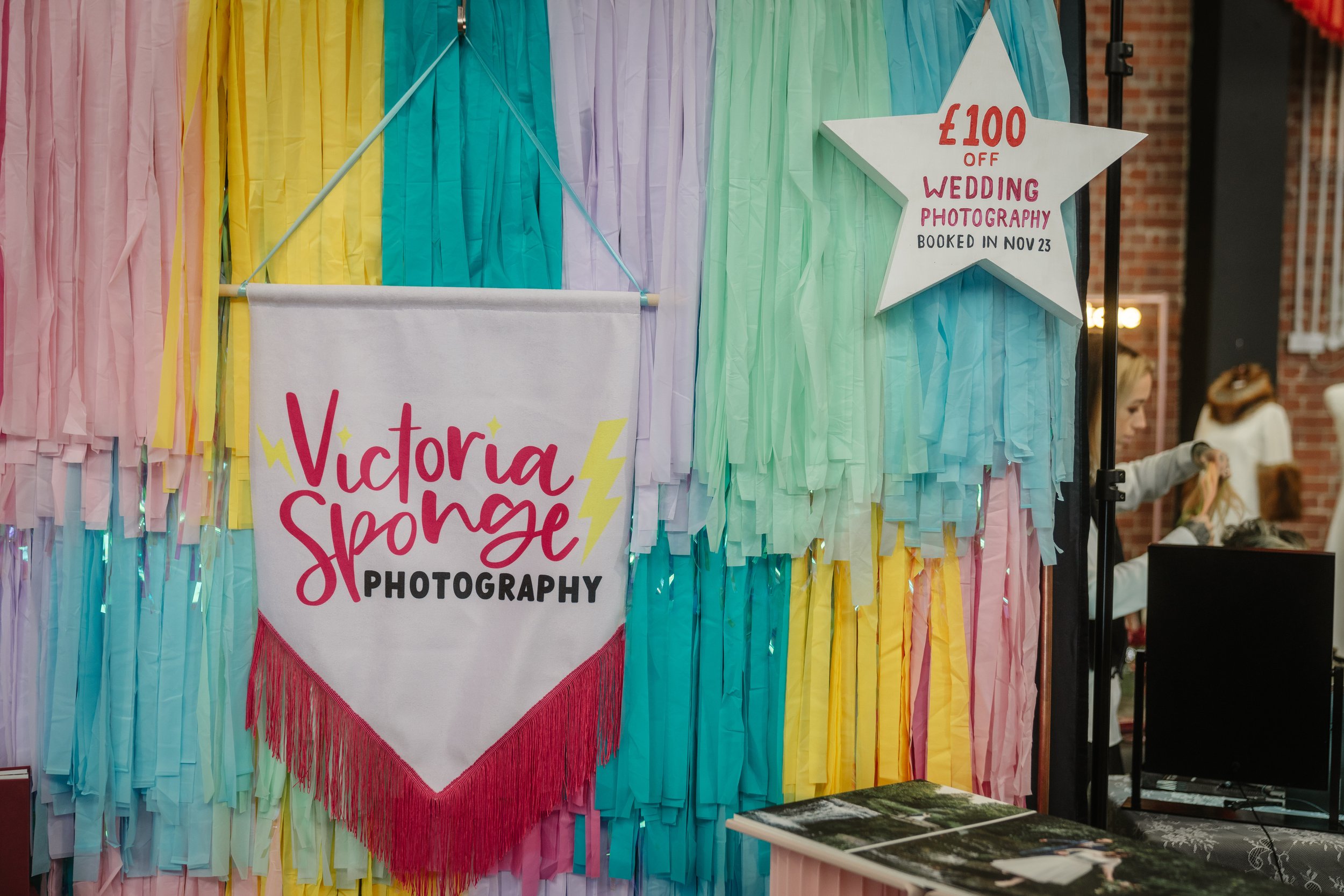  VICTORIA SPONGE wedding photographer’s banner is hung in front of layered multicoloured streamers at their stand at TUWS. 