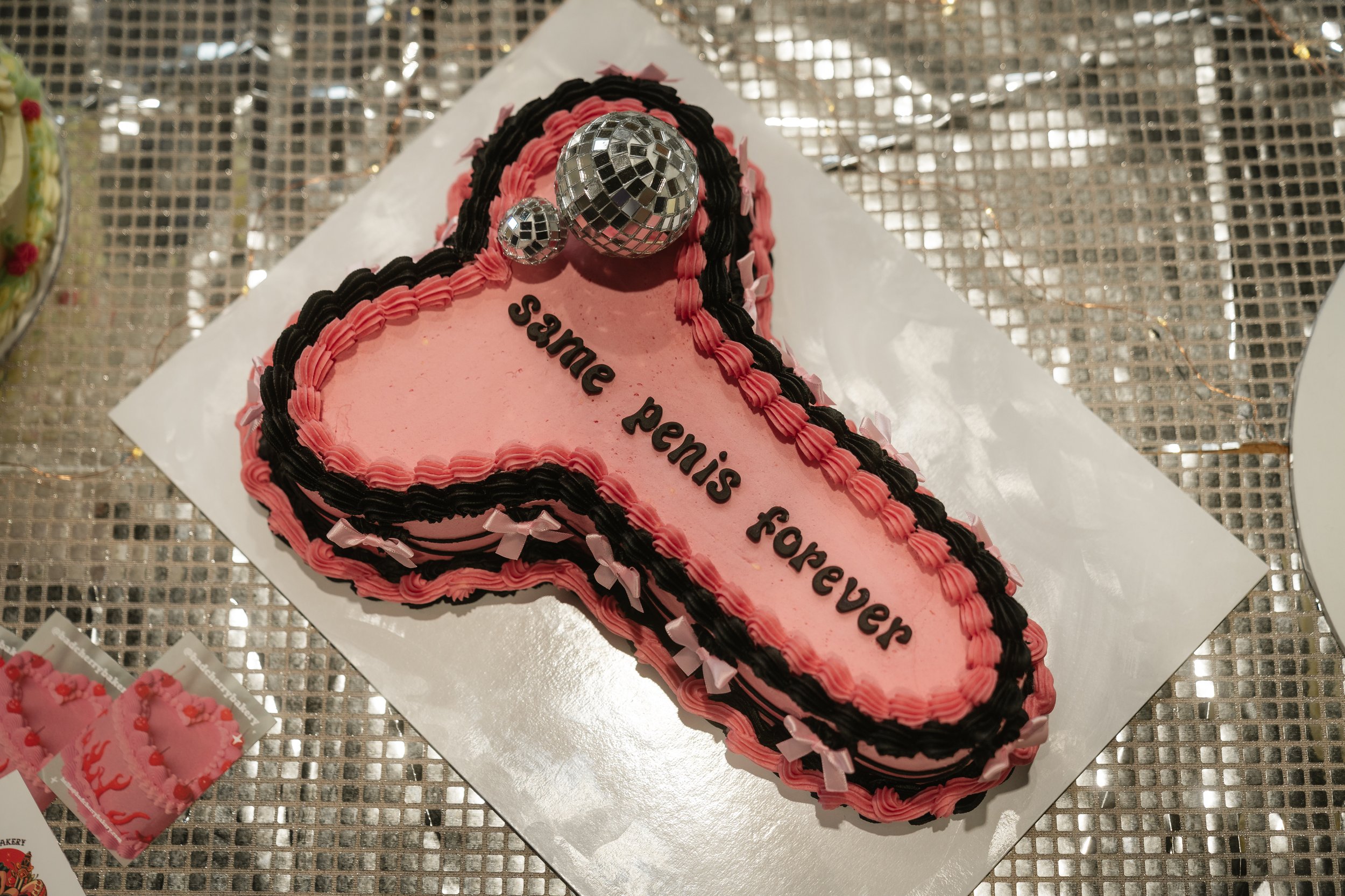  SAME PENIS FOREVER cake with bows around the edge and mini mirror balls on top is displayed on a shiny silver metallic tablecloth. 