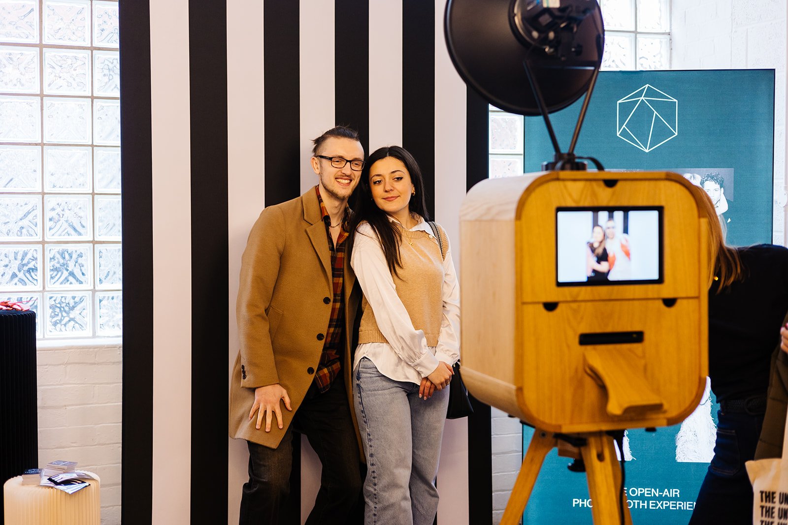  An engaged coupled standing in front of a black and white stripey backdrop and having their photo taken by a wooden photobooth. 