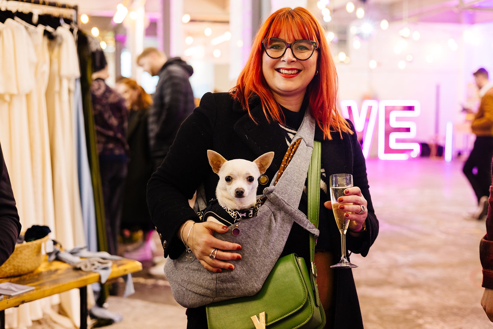  A woman with bright red hair and glasses is holding a glass of champagne and a chihuahua in a sling at The Un-Wedding Show.   