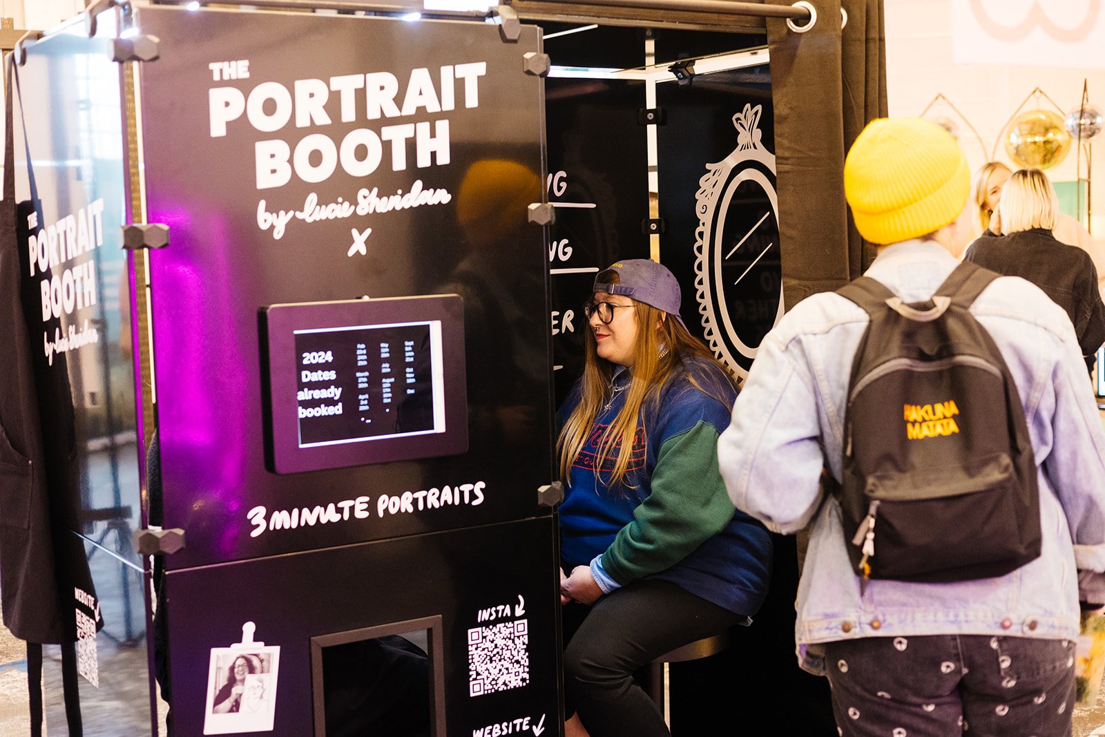  A person with long hair is sitting inside a portrait booth and having their picture drawn. 