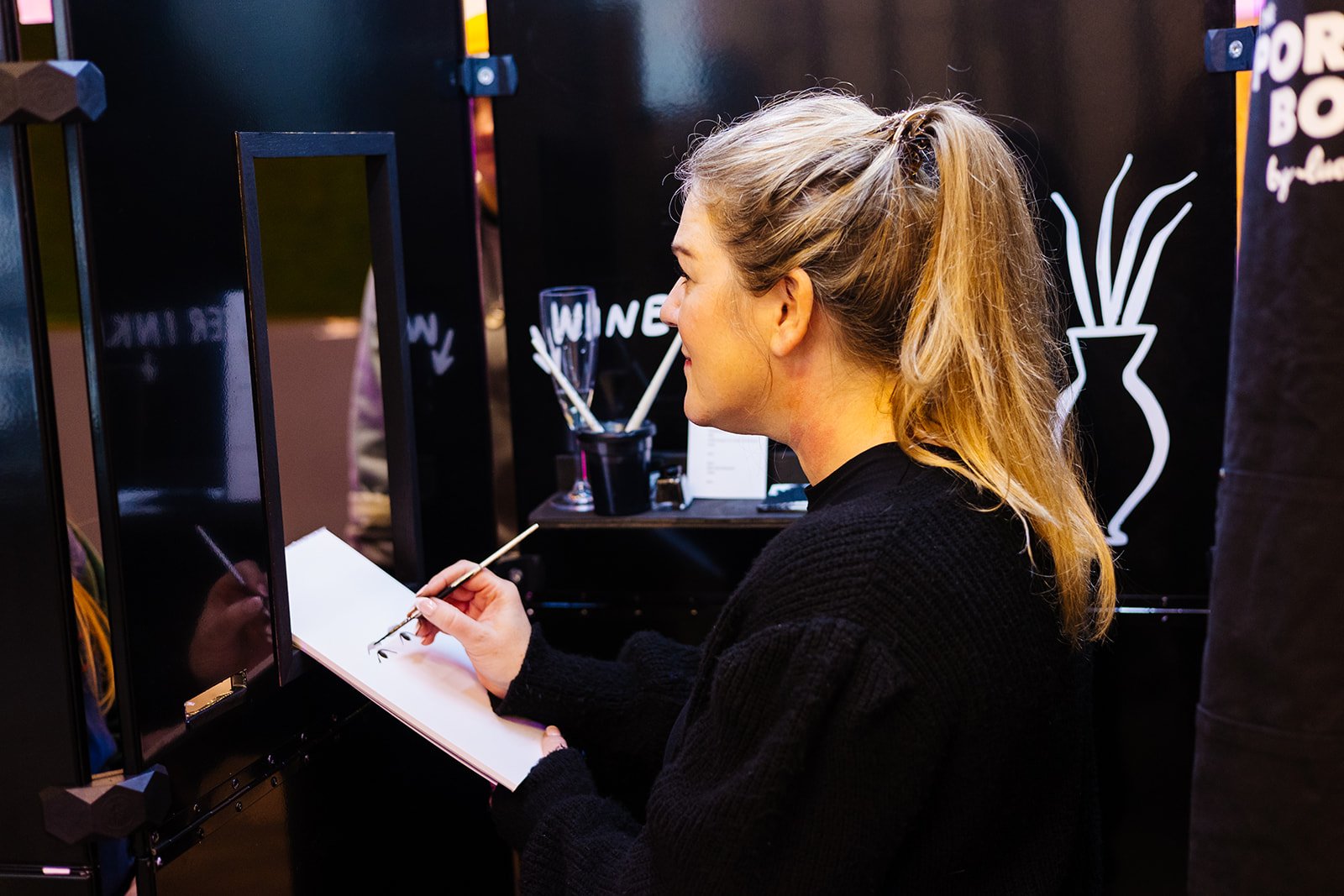  A woman with a long blonde ponytail is drawing someone’s portrait at a wedding fair. 