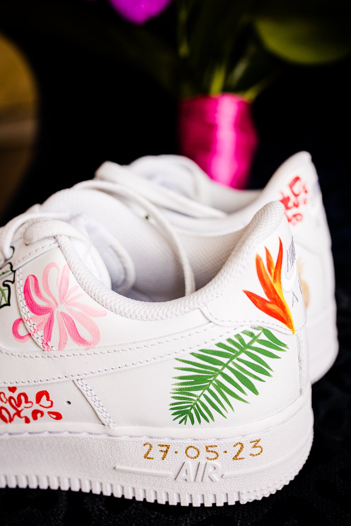  A pair of custom-designed white trainers featuring vibrant hand-painted floral and tropical designs. The shoes display pink hibiscus flowers, green palm leaves, and a bold orange bird-of-paradise flower. Gold numbers "27.05.23" are inscribed near th