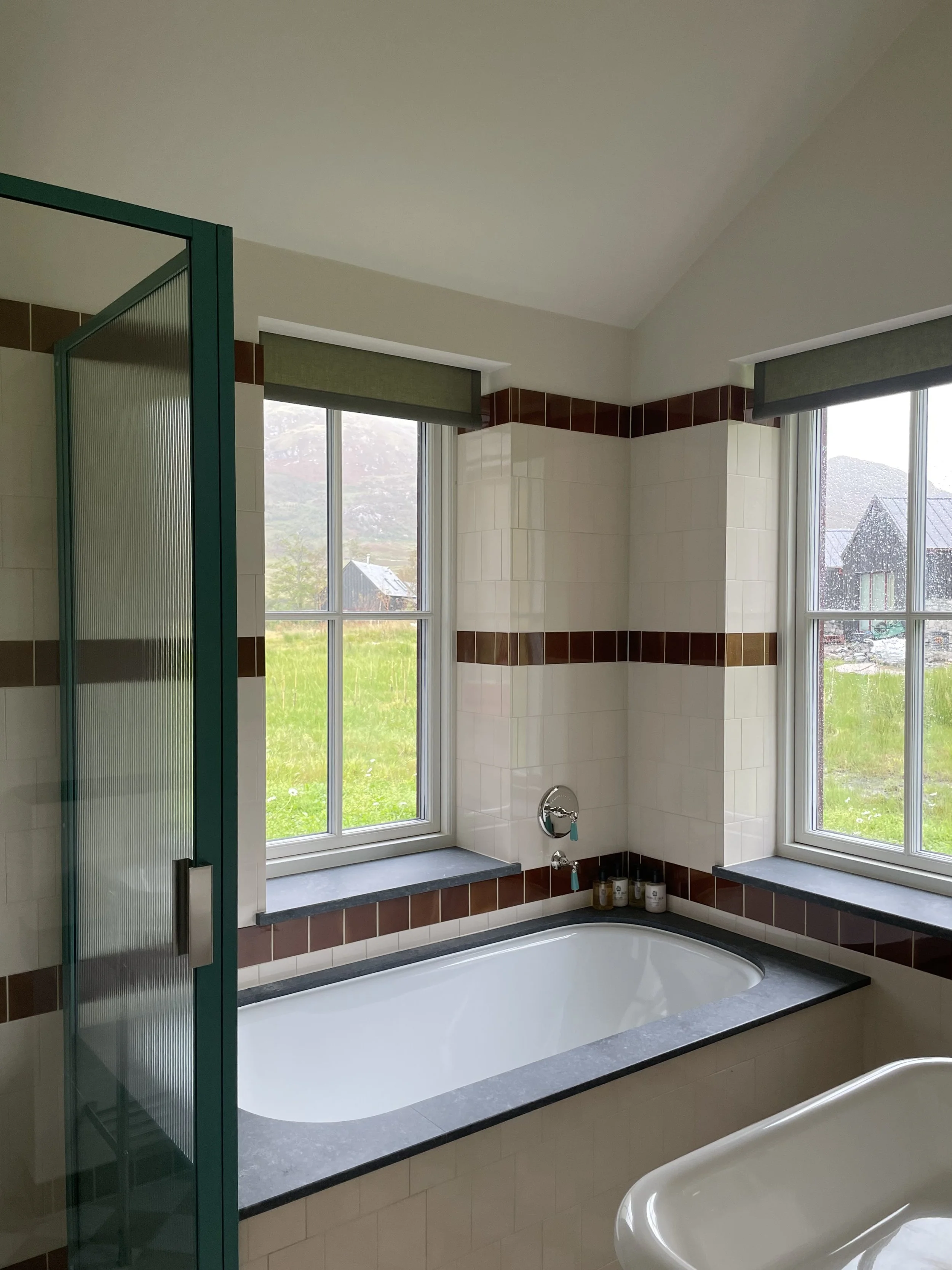 A bathroom with a bathtub next to two windows and a shower enclosure to the left. The bathtub has a dark gray countertop around it, and there are small bottles on the ledge. The bathroom features white tiles with brown border accents and has green window valances.