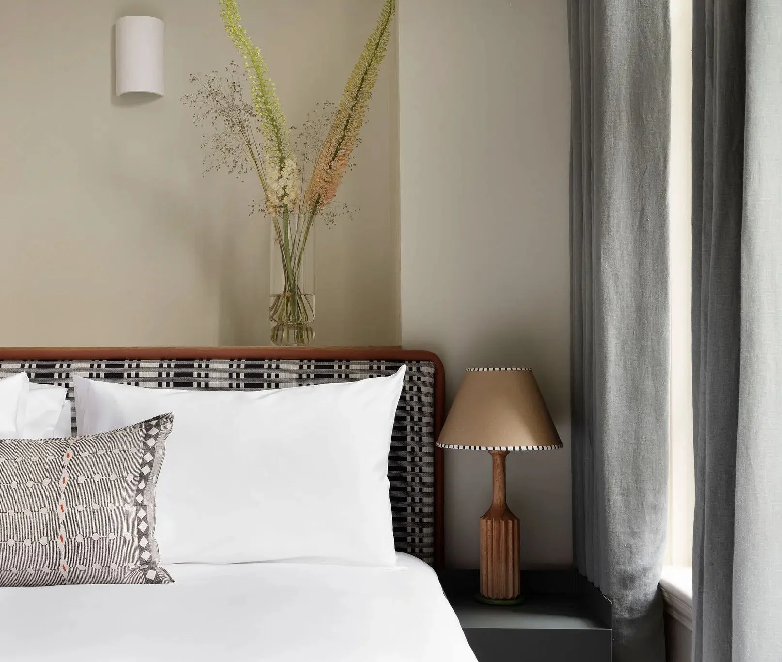 A bedroom with a bed, decorative pillow, bedside lamp, and a vase with tall dried flowers on the wall behind the bed next to curtains.