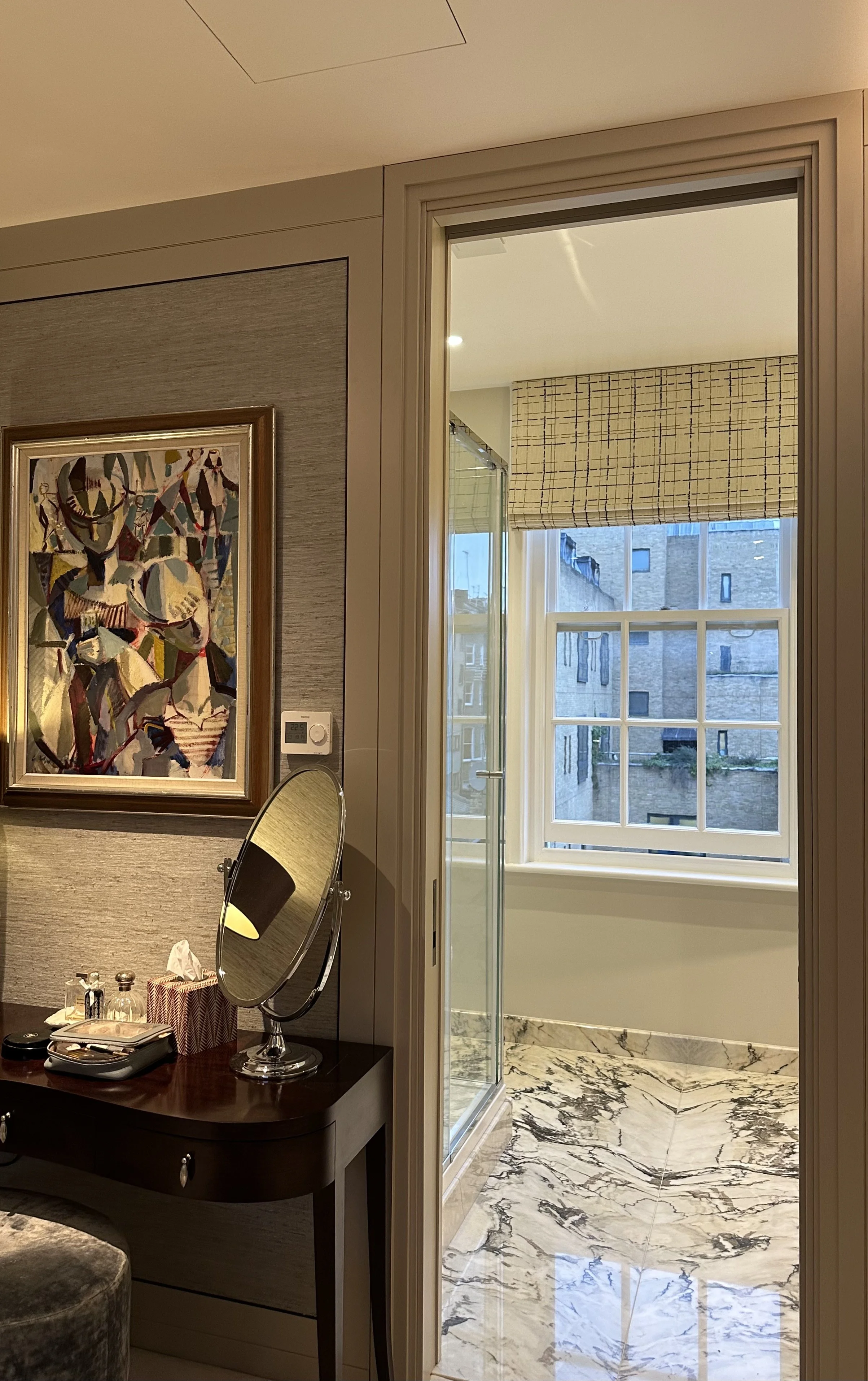 Interior view of a room with a dresser, a mirror, and artwork on the wall, leading to a bright space with large windows and marble flooring outside.