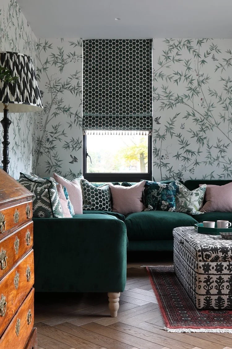 Cozy living room with a dark green velvet sofa, patterned cushions, and a black and white patterned ottoman on a wooden floor. A window with black and white shades, floral and bamboo wallpaper, a wooden dresser, and a large black and white lamp are also visible.