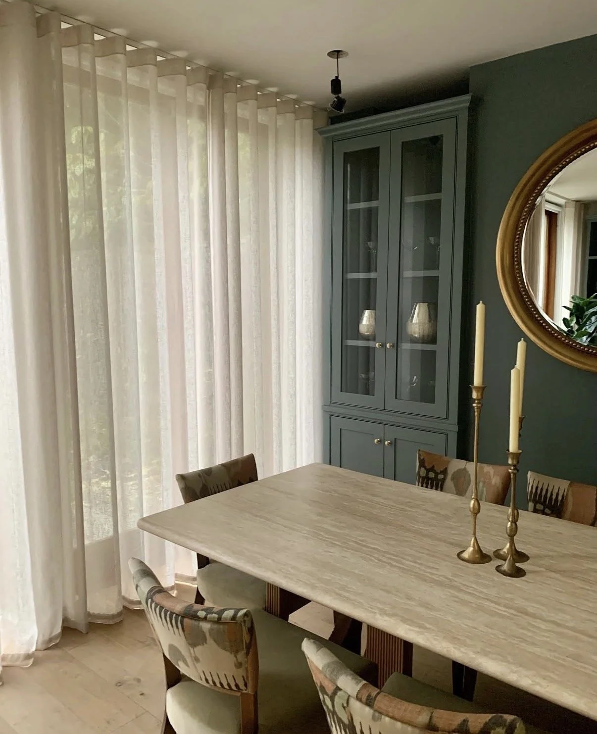 Dining room with a wooden table, upholstered chairs, a tall blue cabinet with glass doors, a round mirror, and two gold candlesticks with candles, with curtains covering a window.