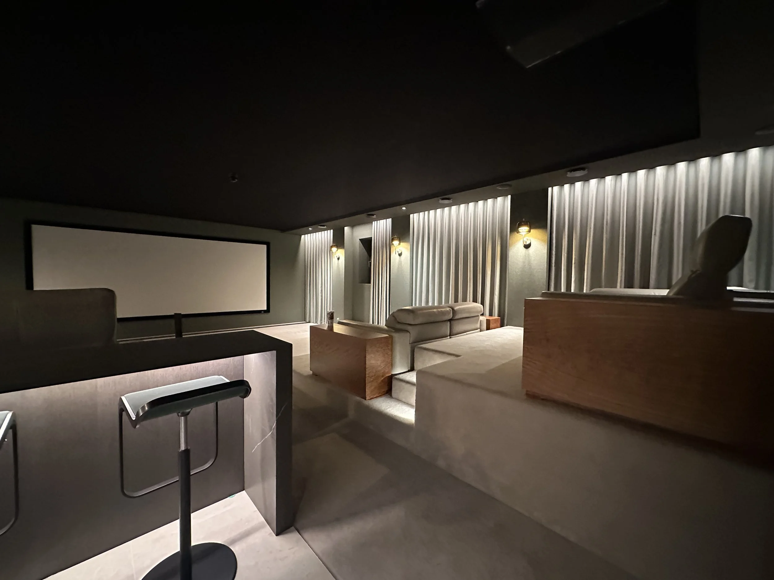 Darkly lit home theater with a large projector screen, beige sofas, and curtains along the back wall.