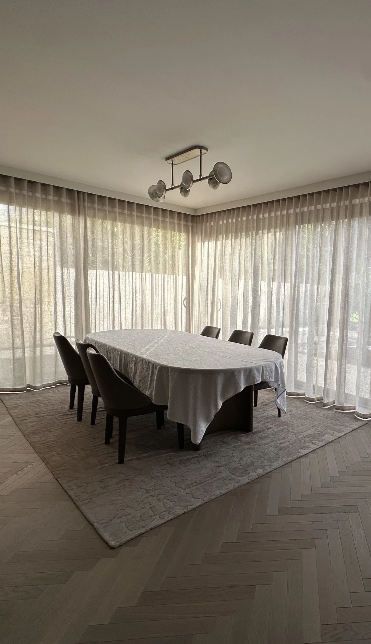 Dining room with a table covered by a white tablecloth, surrounded by six black chairs, in front of large windows with sheer curtains, and a modern ceiling light fixture.