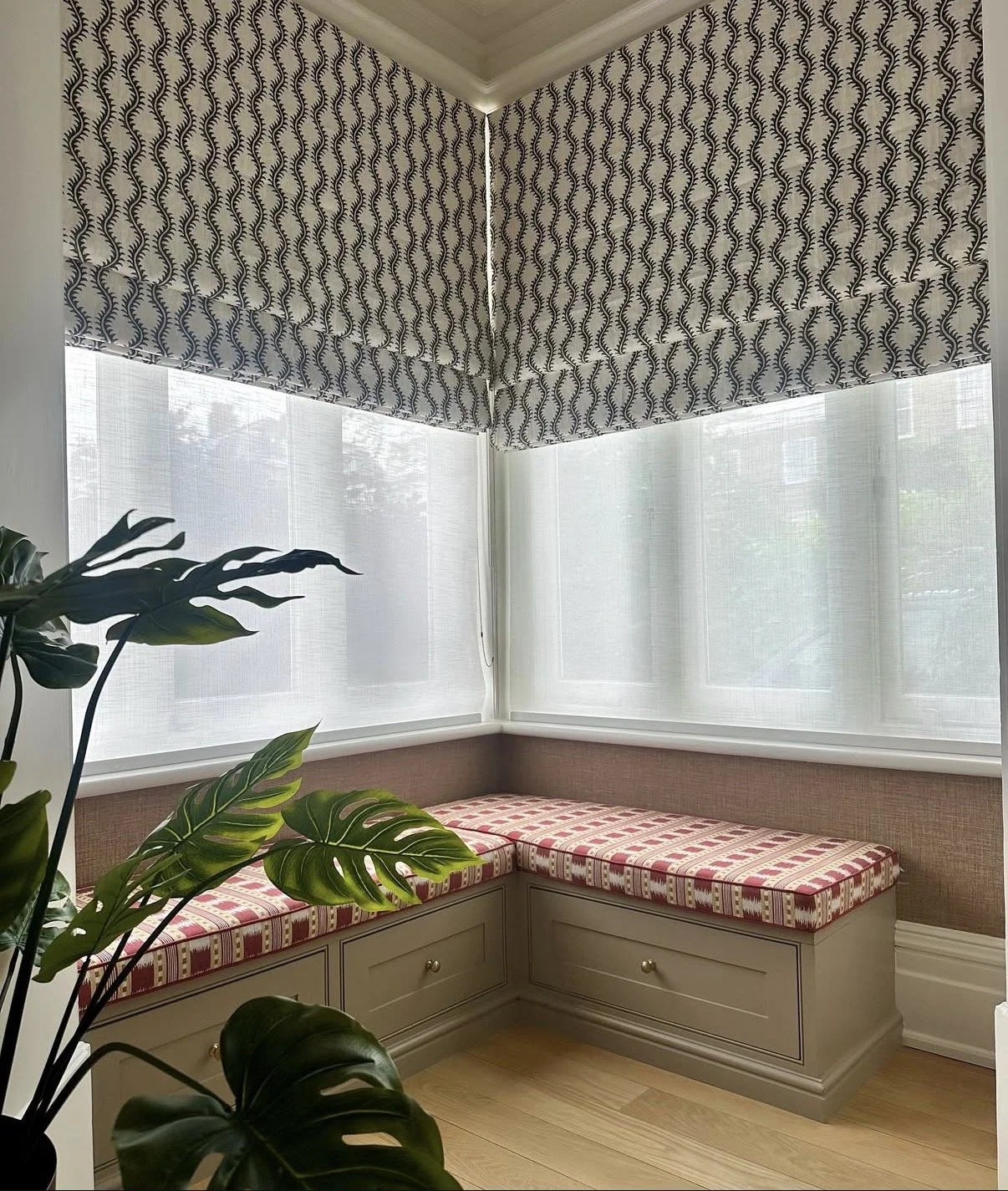 A cozy window nook with built-in storage drawers, cushioned seating with a red checkered pattern, large windows with sheer white blinds, patterned roman shades, a potted plant with large green leaves, and light wood flooring.