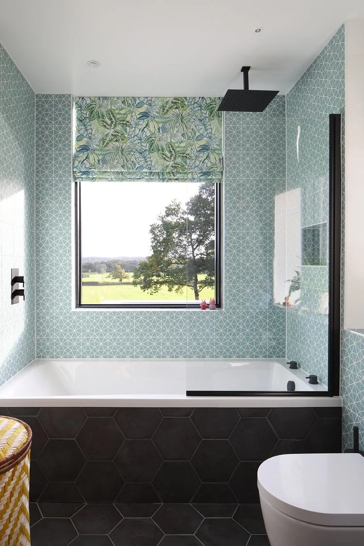 Modern bathroom with a bathtub beneath a window showing a scenic outdoor view, featuring a tropical-themed window shade, black fixtures, a walk-in shower with a black shower head, geometric patterned light blue tiles on the walls, and black hexagonal tiles on the floor.