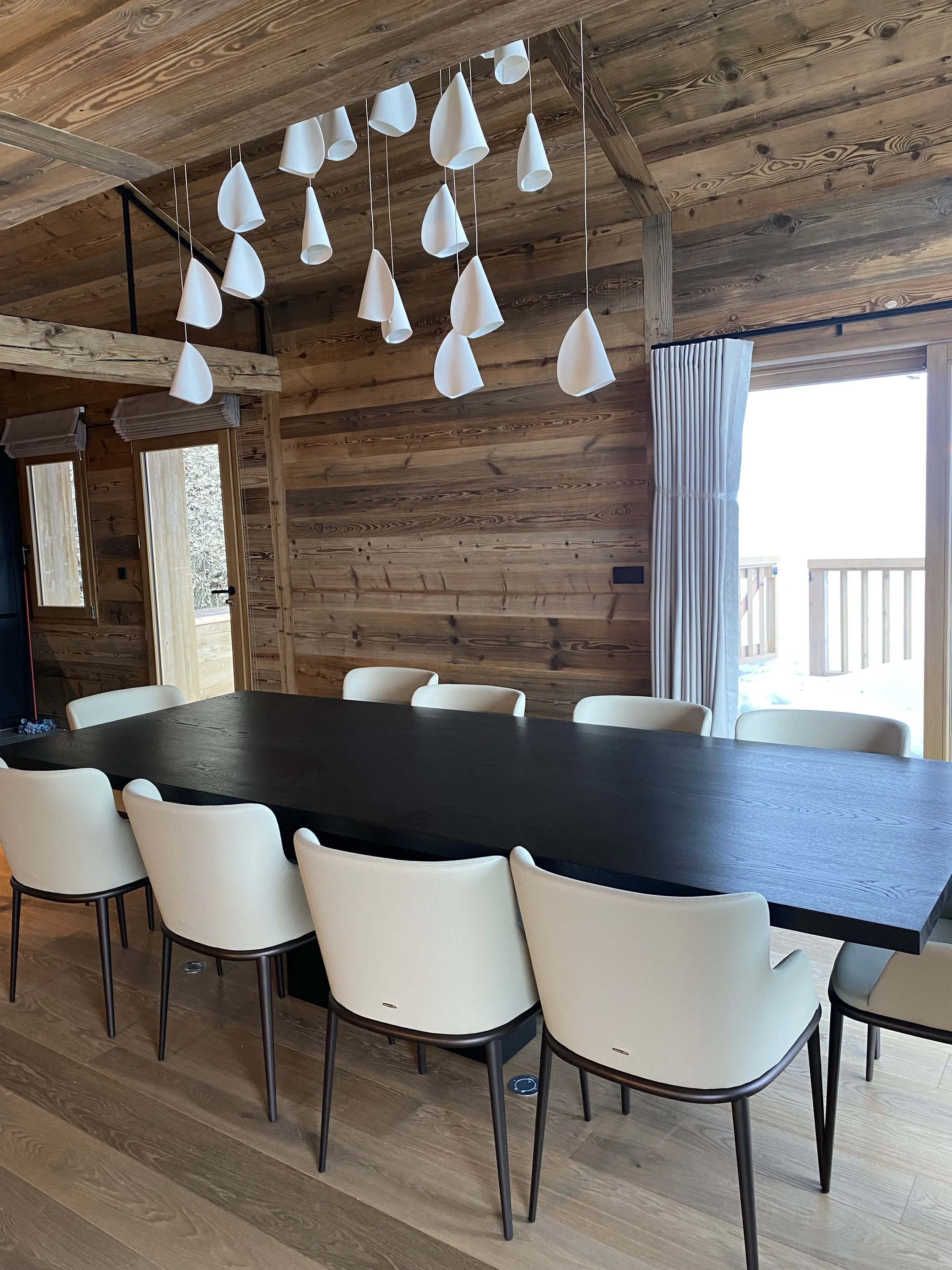 Interior of a dining room with a long dark wood table, white chairs, wood-paneled walls, a sliding glass door with a curtain, and a modern hanging light fixture with white cone-shaped shades.