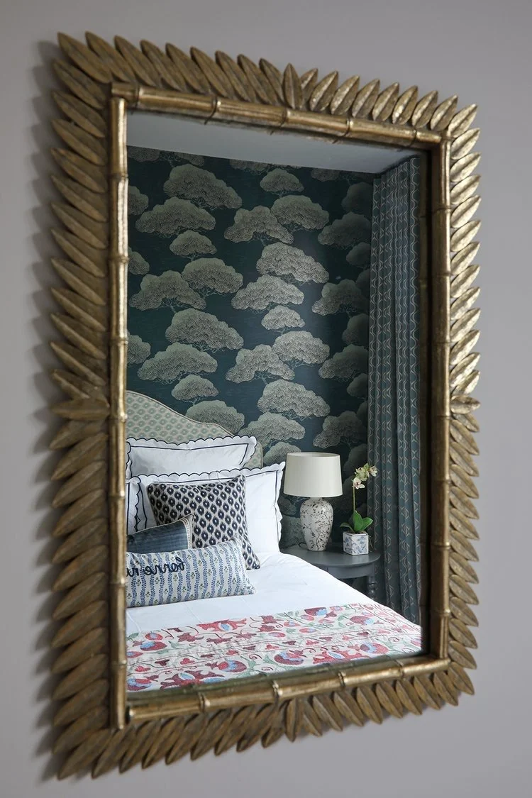 Reflection of a bedroom with a bed, patterned pillows, a bedside lamp, and floral wall decor, seen in a large decorative gold framed mirror.