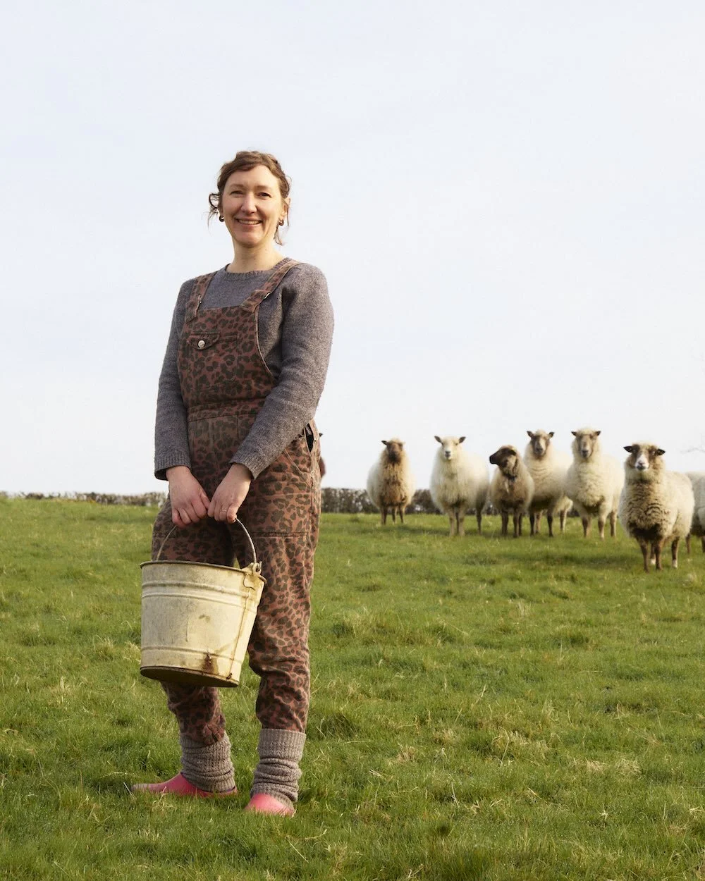 Xanthe standing holding a metal bucket with her sheep behind her.