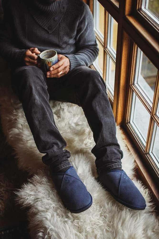 Man wearing sheepskin slippers, sitting on a sheepskin rug, holding a mug of tea.