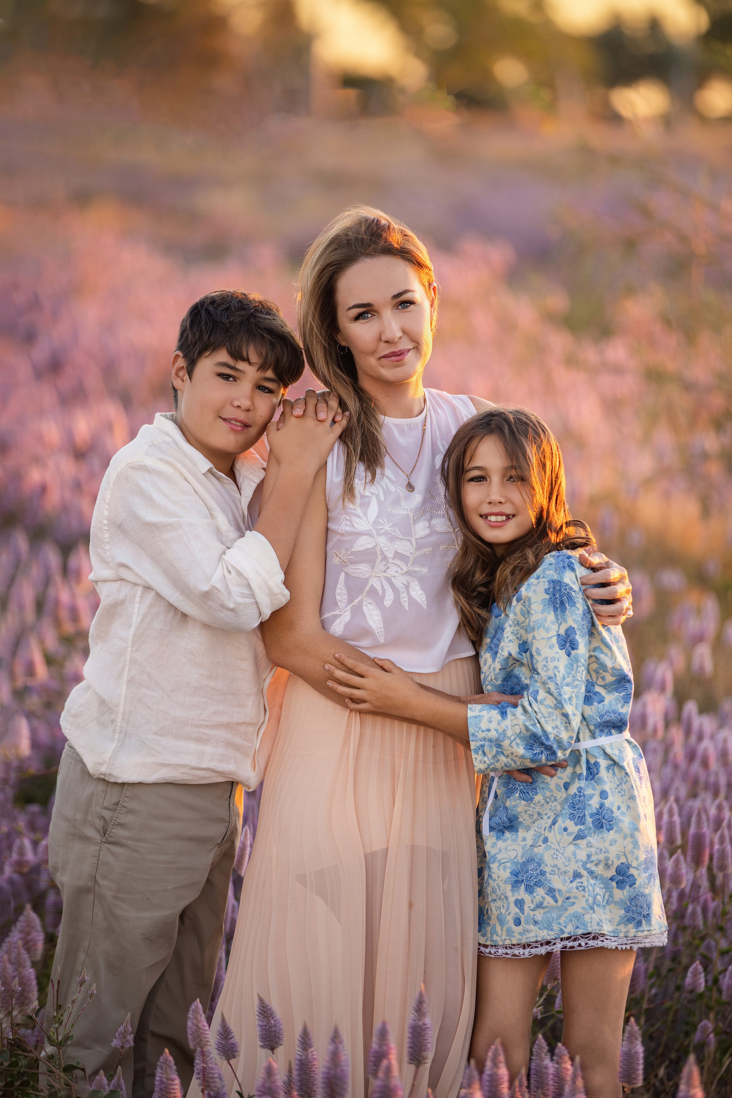 A woman and two children stand together in a field of purple flowers at sunset, smiling at the camera.  Outdoor family photoshoot, Karratha photographer