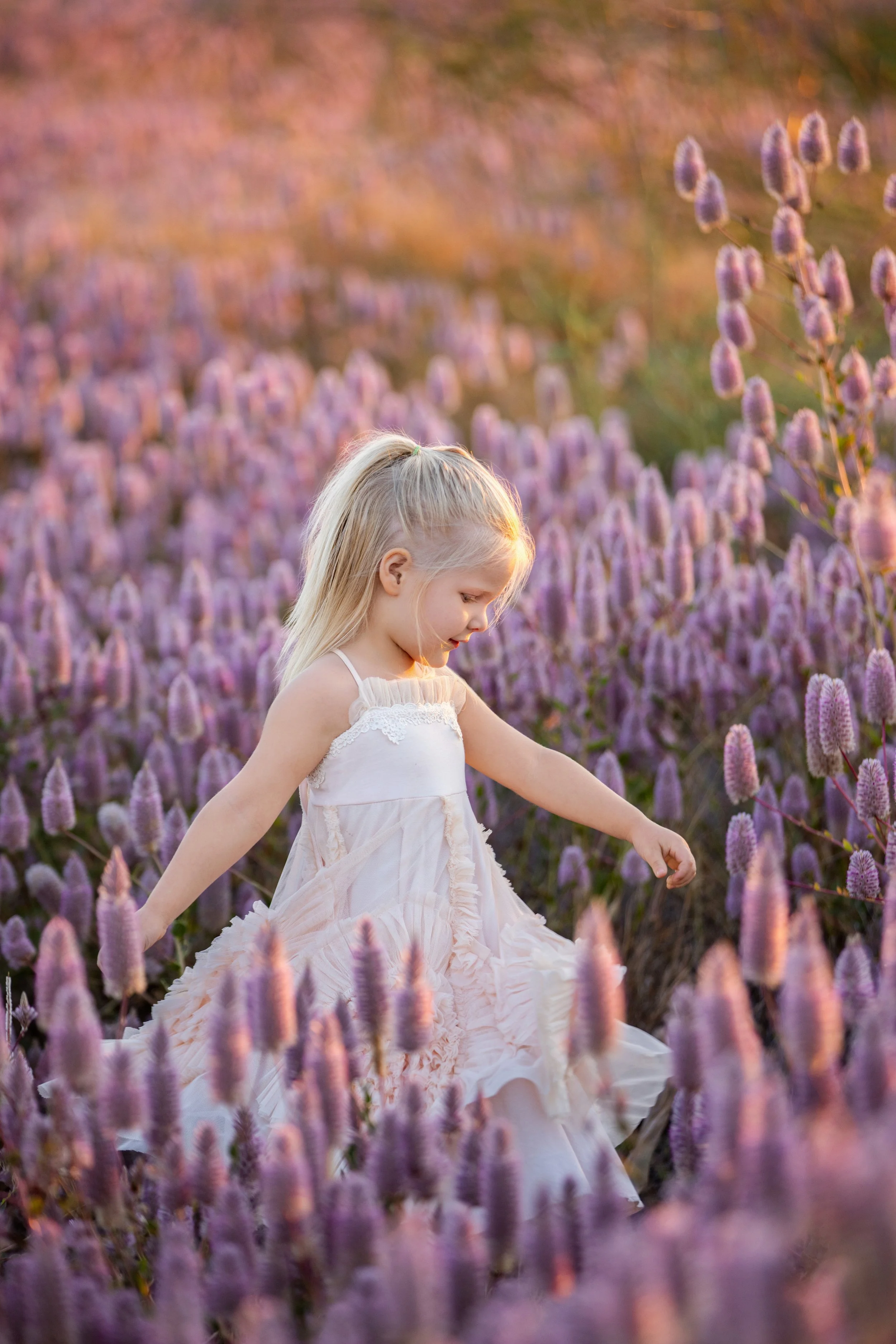 A young girl with long blonde hair in a braid, wearing a white dress, is walking through a field of purple flowers during sunset.