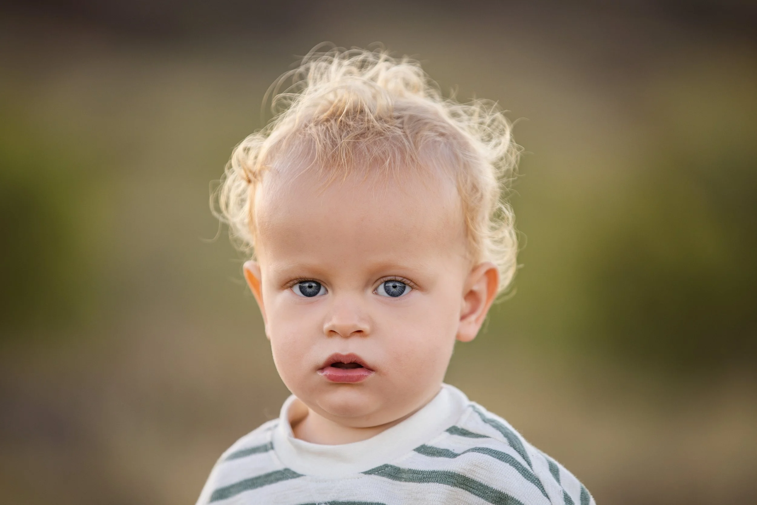 A young child with curly blonde hair and blue eyes wearing a striped shirt, looking slightly to the side with a serious expression.