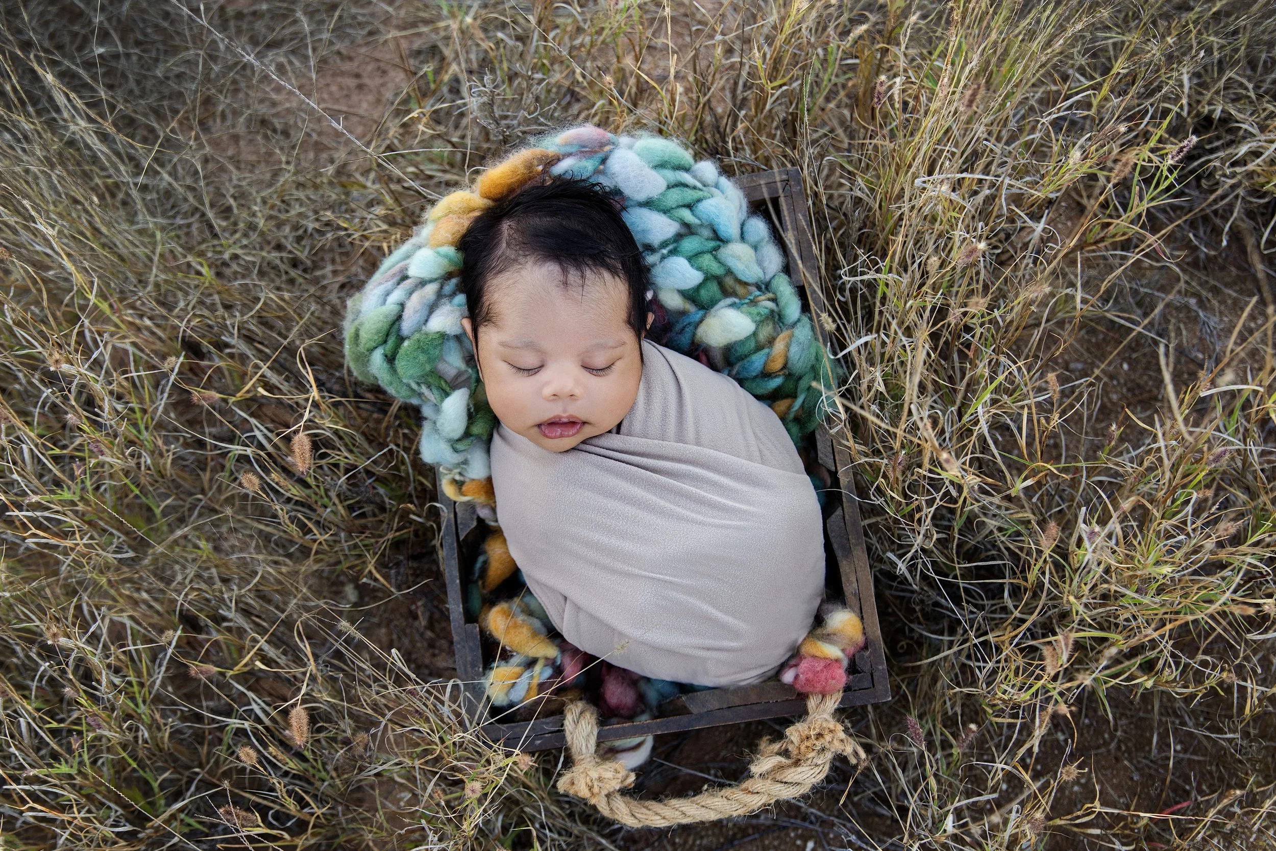 A young child with short black hair is sleeping outdoors on a wooden platform, wrapped in a multicolored blanket, surrounded by dry grass. Outdoor newborn photoshoot, Karratha photographer