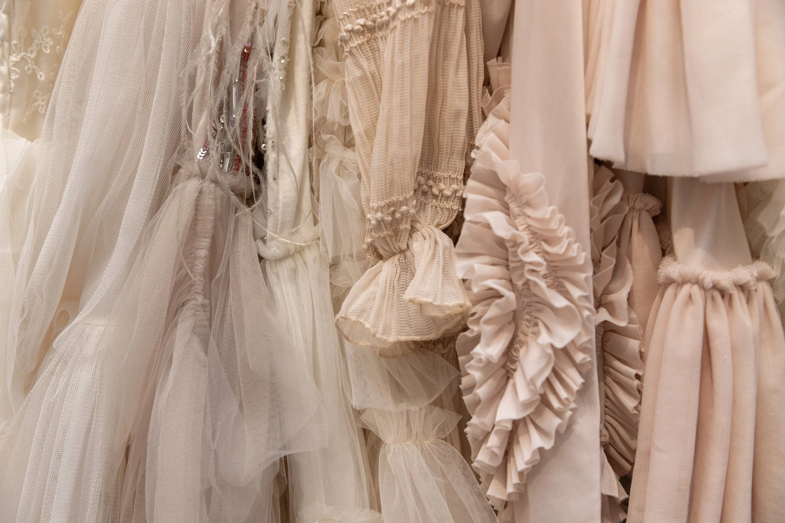 Photography client wardrobe. A row of beige and white dresses made of different fabrics, including tulle, chiffon, and lace, hanging on a rack in a clothing store.
