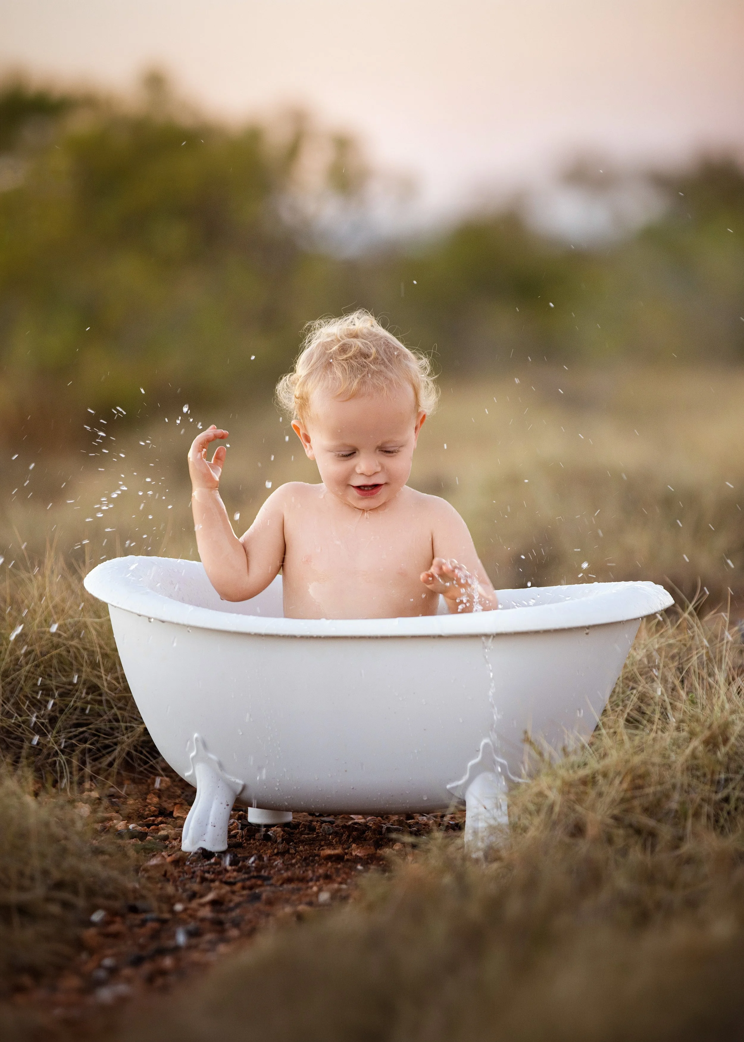 A young child with curly blonde hair playing in a white porcelain bathtub outdoors on a grassy area, splashing water, during sunset or late afternoon.
