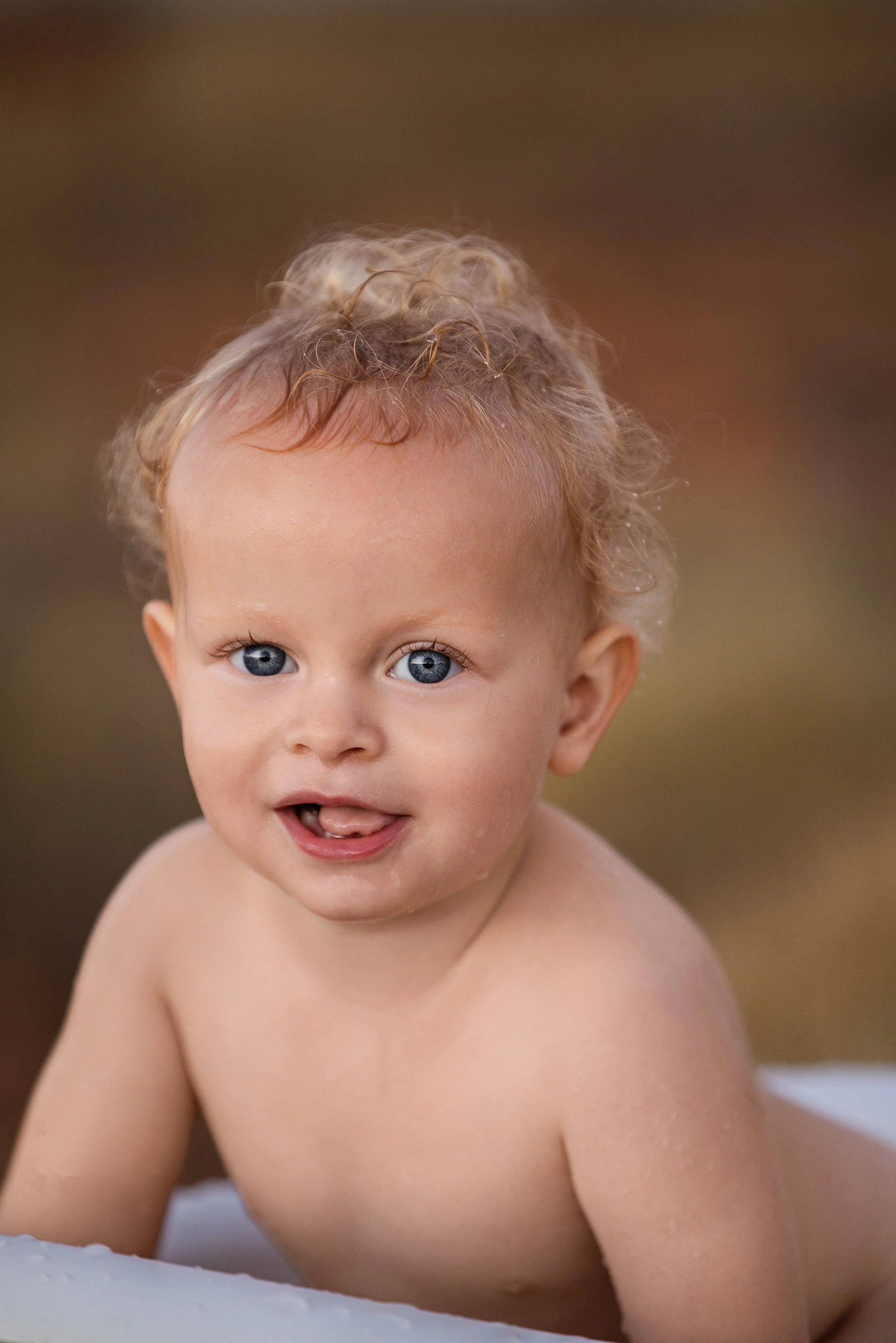 A young child with curly blonde hair and blue eyes looking up and smiling, with a blurred brown background.  Outdoor family photoshoot, Karratha photographer