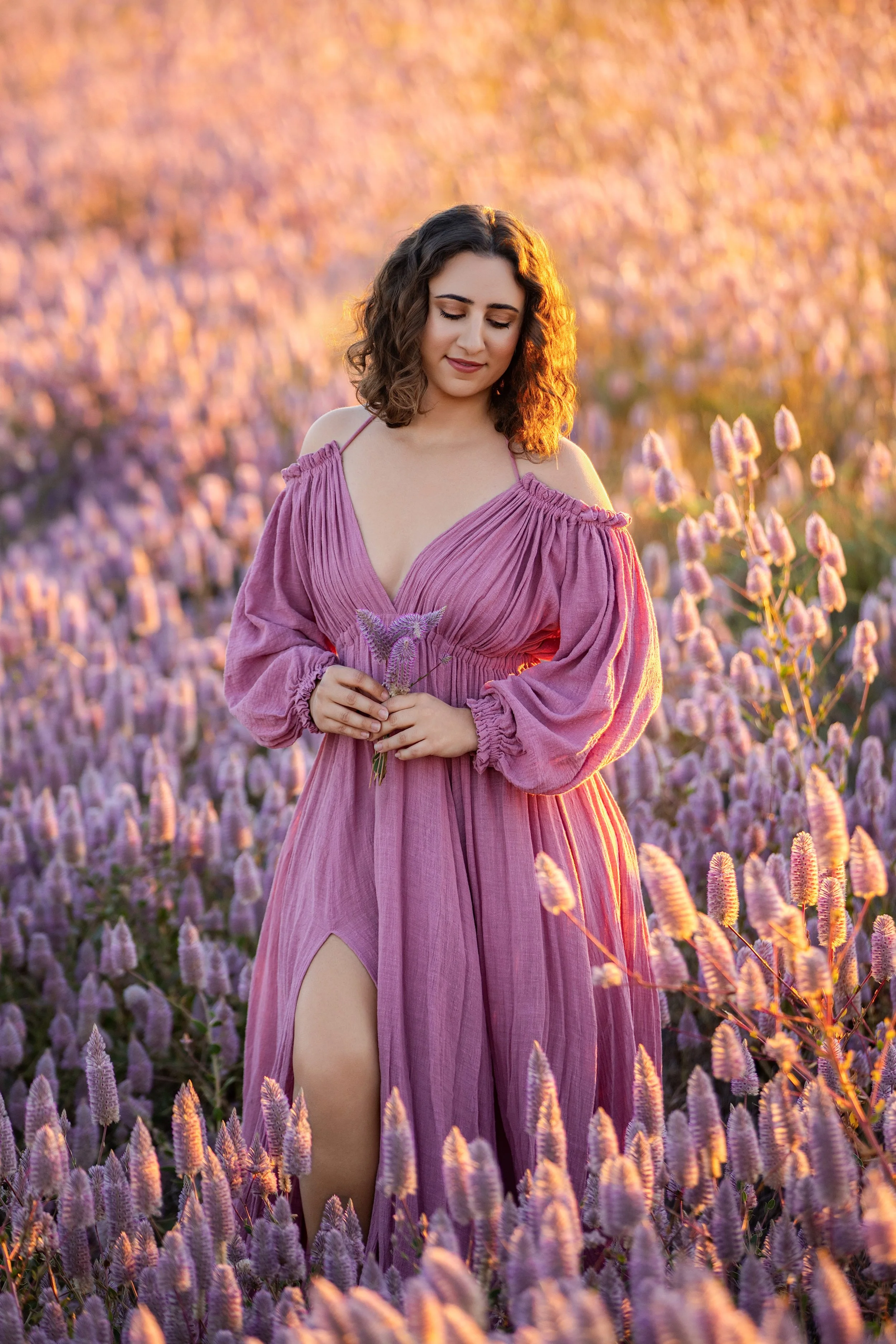 A woman in a pink dress standing in a lavender field during sunset, holding lavender flowers, with her eyes closed and a gentle expression on her face.