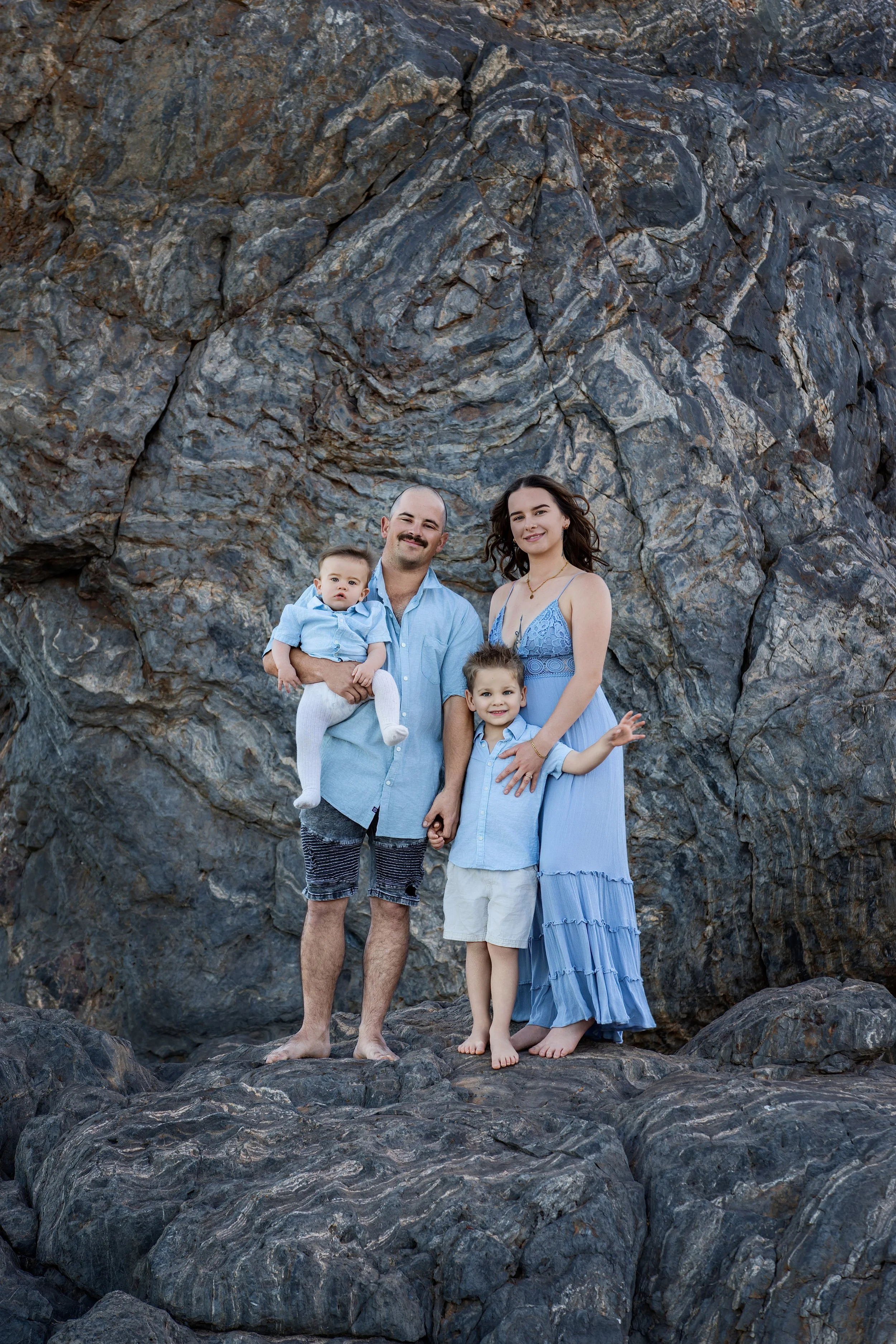 A family of four standing on rocks in front of a large rocky cliff. The father is holding a young boy, and the mother is standing with a young boy. family photography, photographer, photoshoot