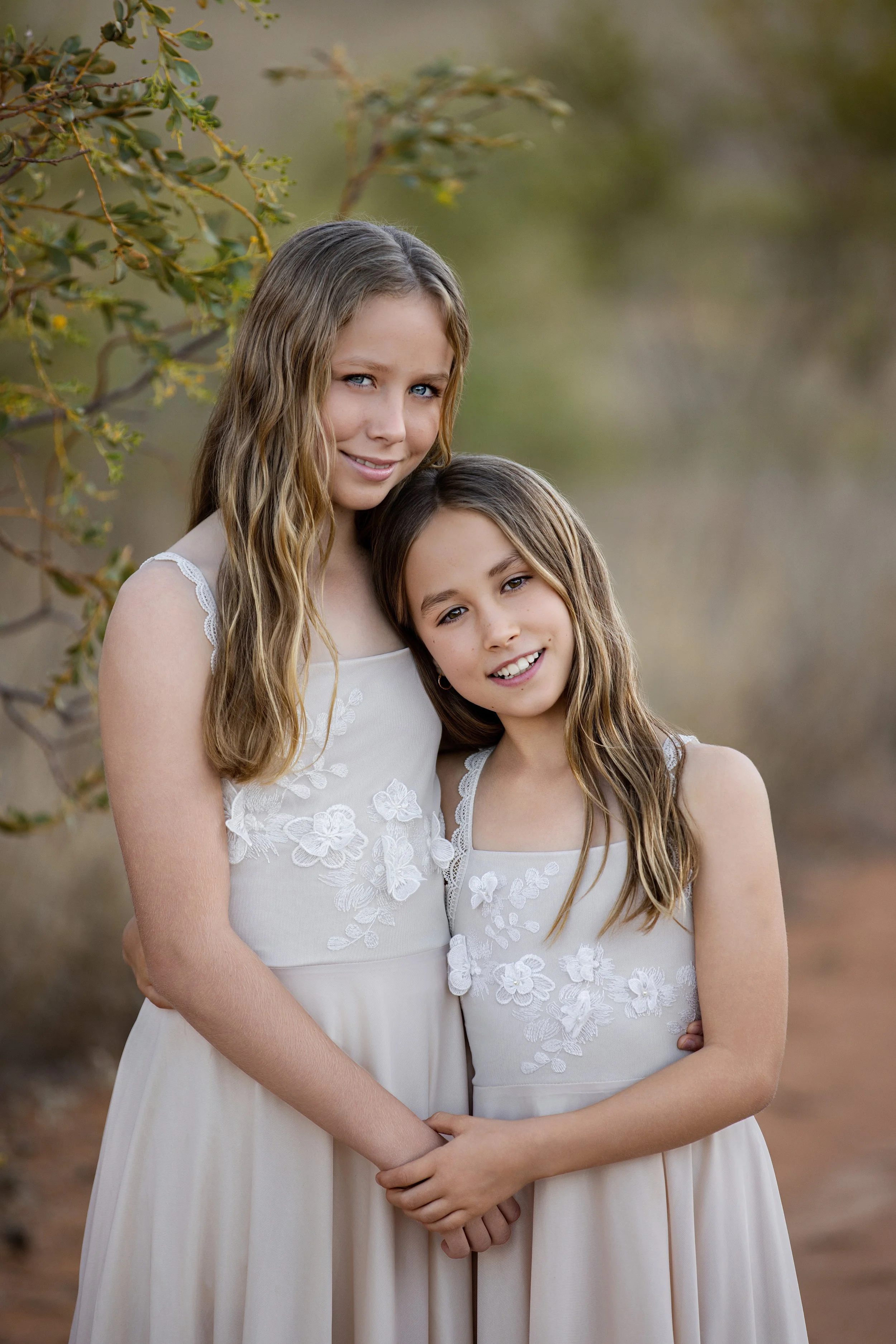 Two young girls in white dresses with floral embroidery standing outdoors, smiling, with trees and foliage in the background.