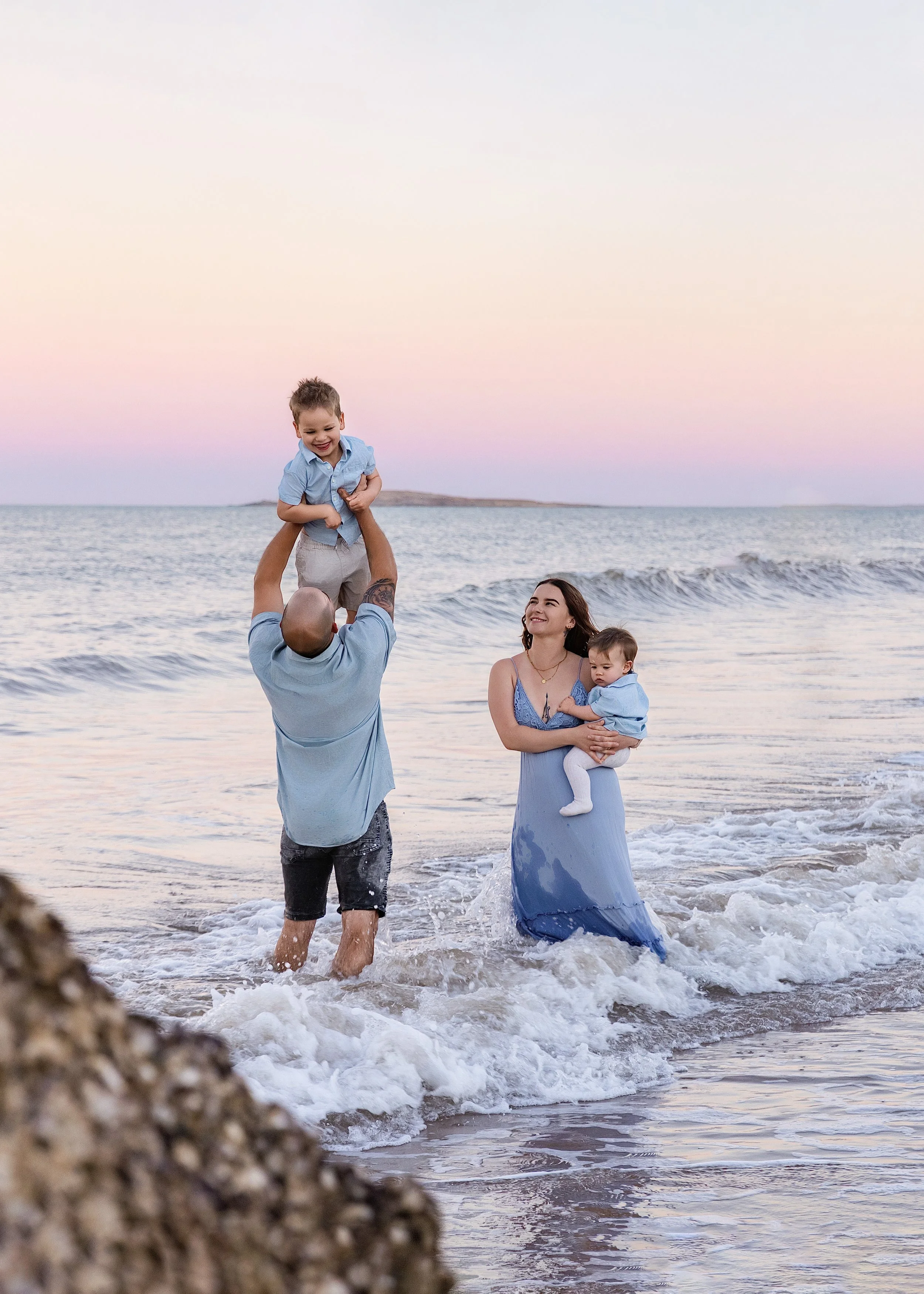 A family on the beach, with a man lifting a boy, a woman holding a baby, and the ocean in the background during sunset.  Outdoor family photoshoot, Karratha photographer