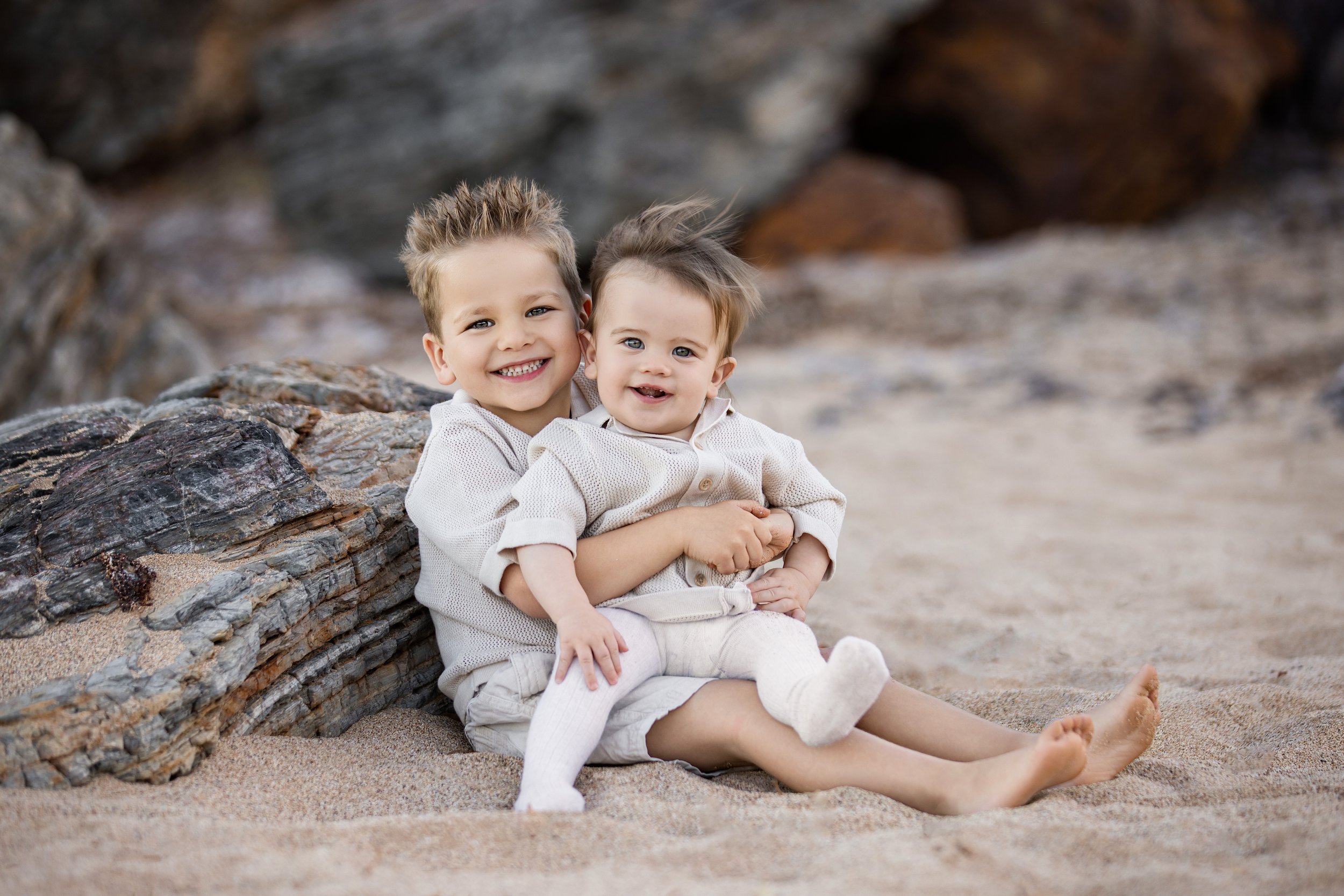 Two young boys sitting on the sand near rocks at the beach, smiling and embracing each other.  Outdoor family photoshoot, Karratha photographer