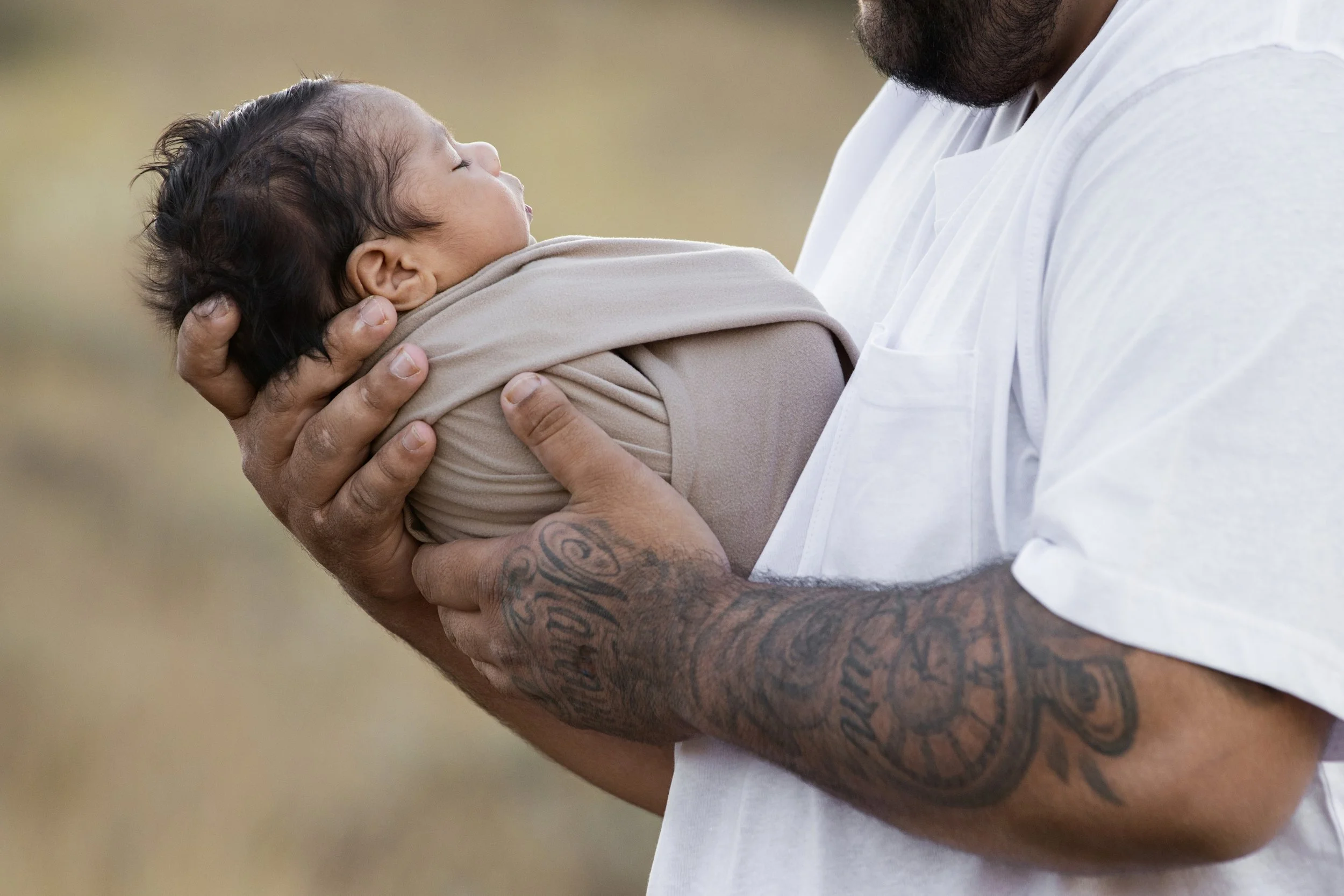 A person with tattoos on their arm holds a sleeping infant wrapped in a beige blanket, close to their chest. Outdoor newborn photoshoot, Karratha photographer